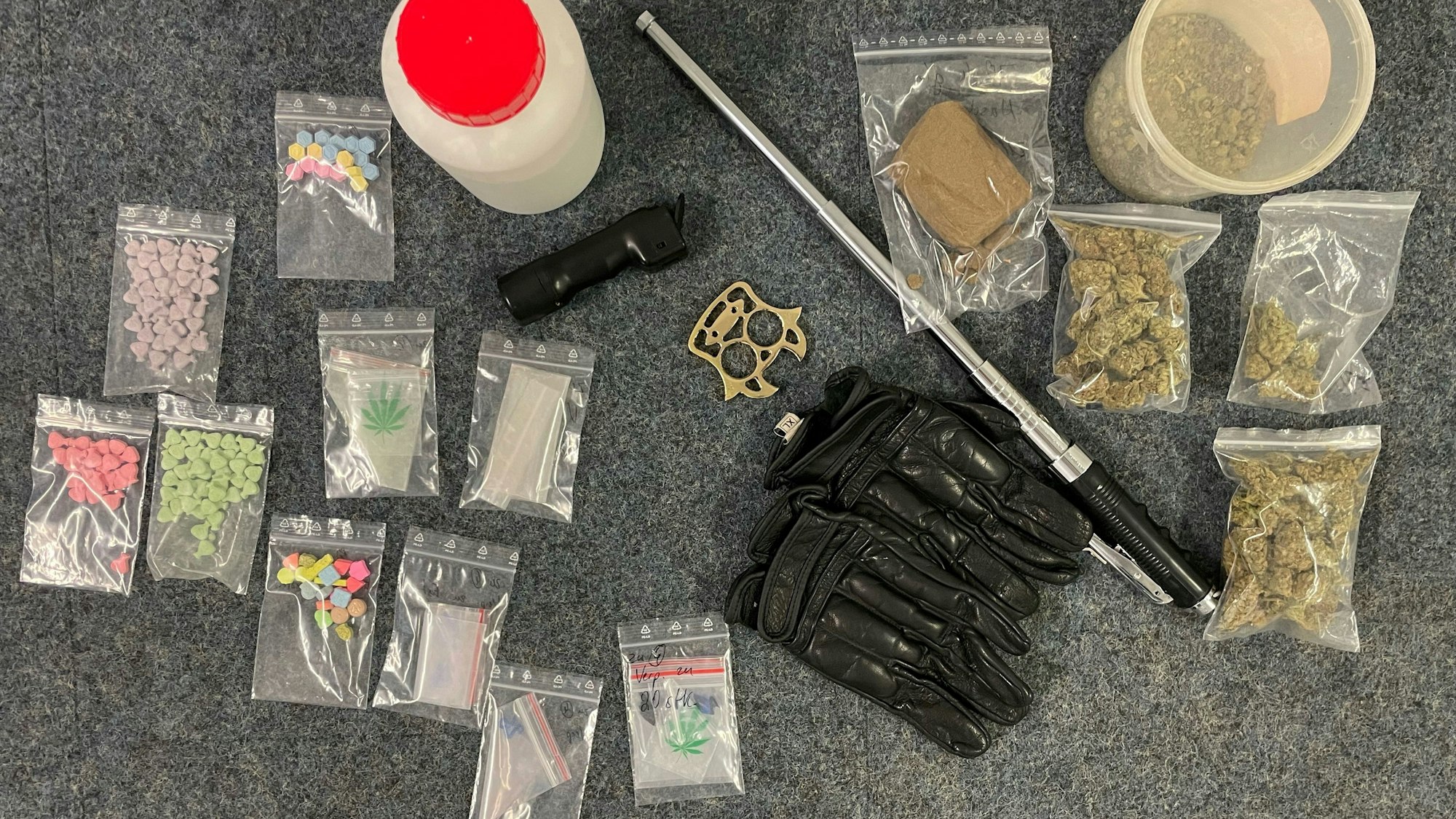 In kleinen Tütchen befinden sich diverse Drogen, dazwischen liegen Waffen wie ein Teleskopschlagstock.
