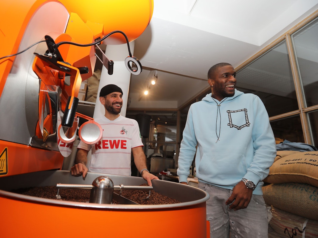 Anthony Modeste und Rafet Aydogdu stehen zusammen an einer Kaffee-Röstmaschine.