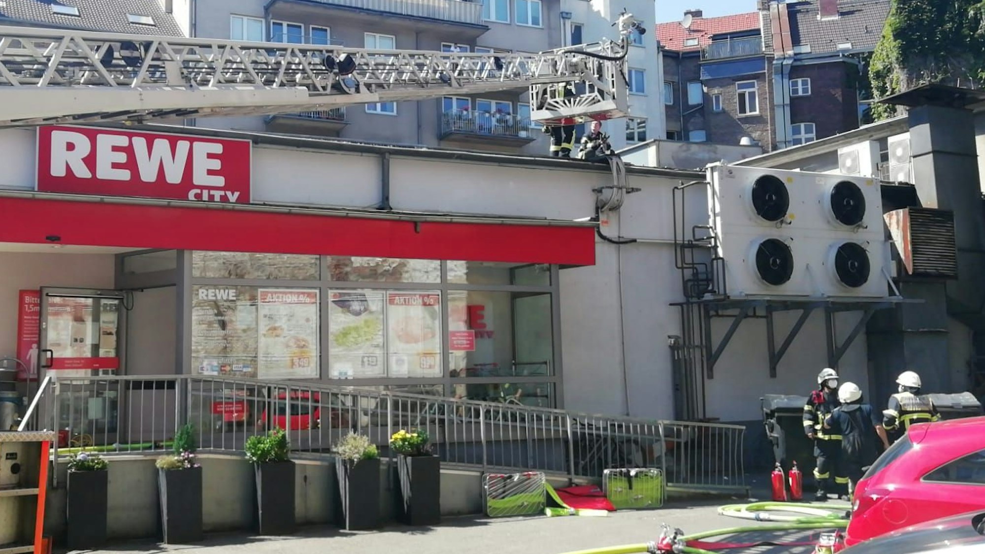 Ein Einsatzfahrzeug mit Drehleiter steht vor einem Rewe-Markt, auf dessenDach sich mehrere Feuerwehrmänner befinden.