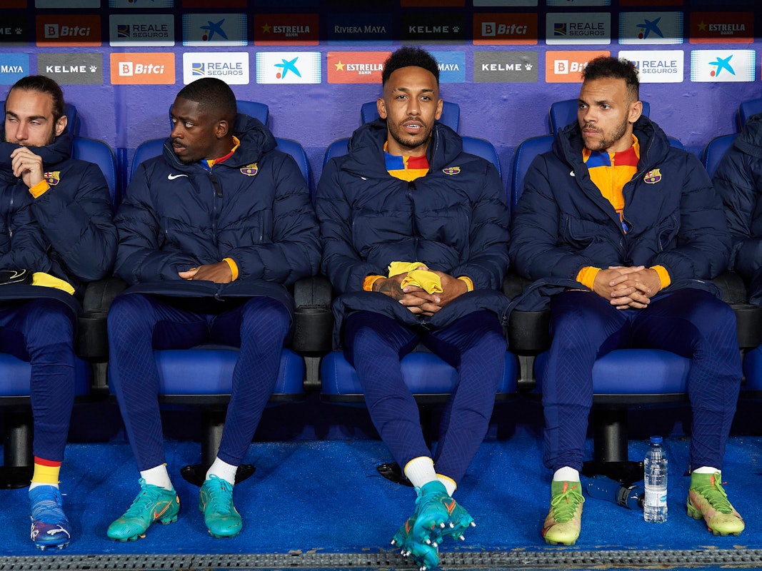 Ousmane Dembele, Pierre-Emerick Aubameyang und Martin Braithwaite (von l. nach r.) vom FC Barcelona sitzen mit wärmenden Jacken auf der Bank.