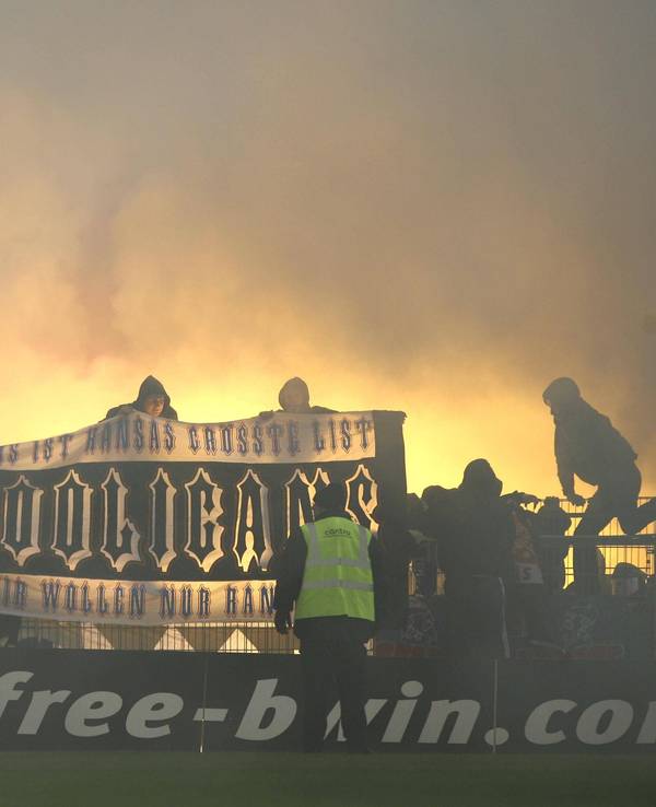 Hansa-Rostock-Fans zeigen auf dem Zaun des Gästeblocks ein Banner, im Hintergrund brennt Pyrotechnik.