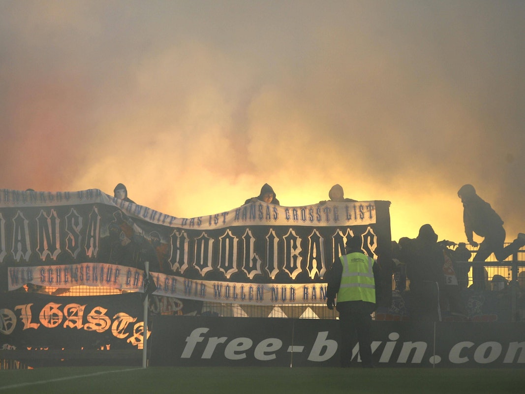 Hansa-Rostock-Fans zeigen auf dem Zaun des Gästeblocks ein Banner, im Hintergrund brennt Pyrotechnik.