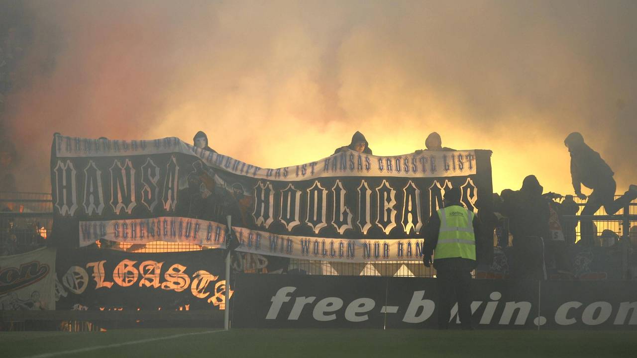 Hansa-Rostock-Fans zeigen auf dem Zaun des Gästeblocks ein Banner, im Hintergrund brennt Pyrotechnik.