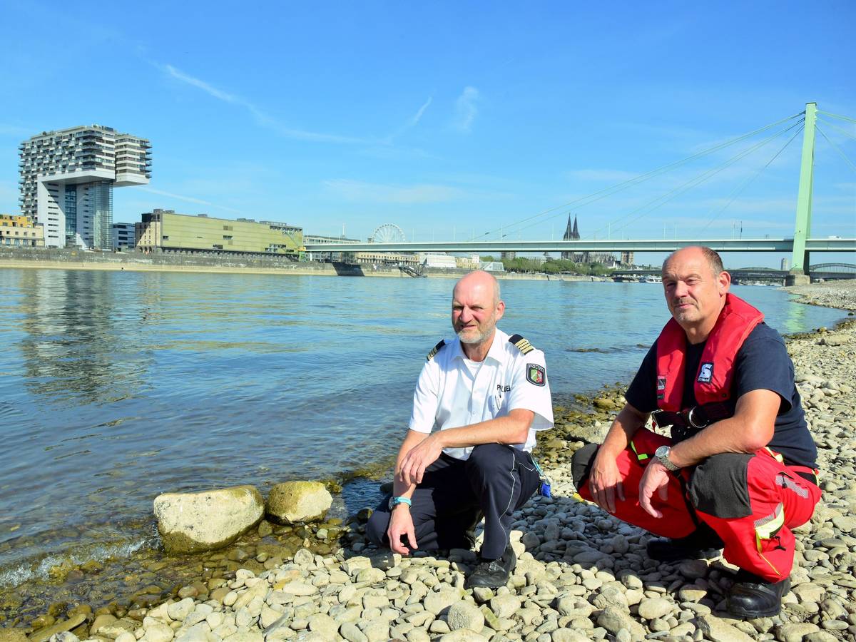 Zwei Kölner Experten hocken direkt am Rheinufer und blicken in die Kamera. Die Gefahren am Rhein sind größer als je zuvor, sagen sie.