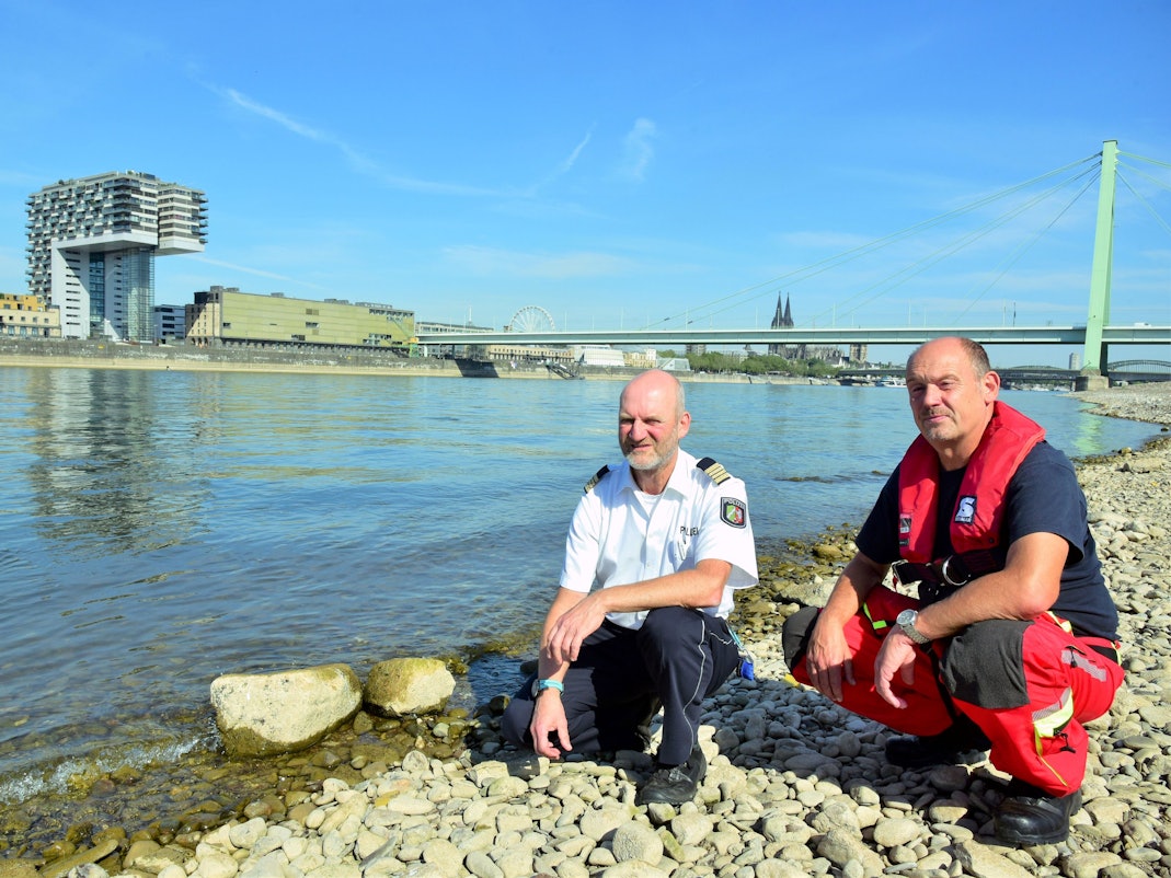 Zwei Kölner Experten hocken direkt am Rheinufer und blicken in die Kamera. Die Gefahren am Rhein sind größer als je zuvor, sagen sie.