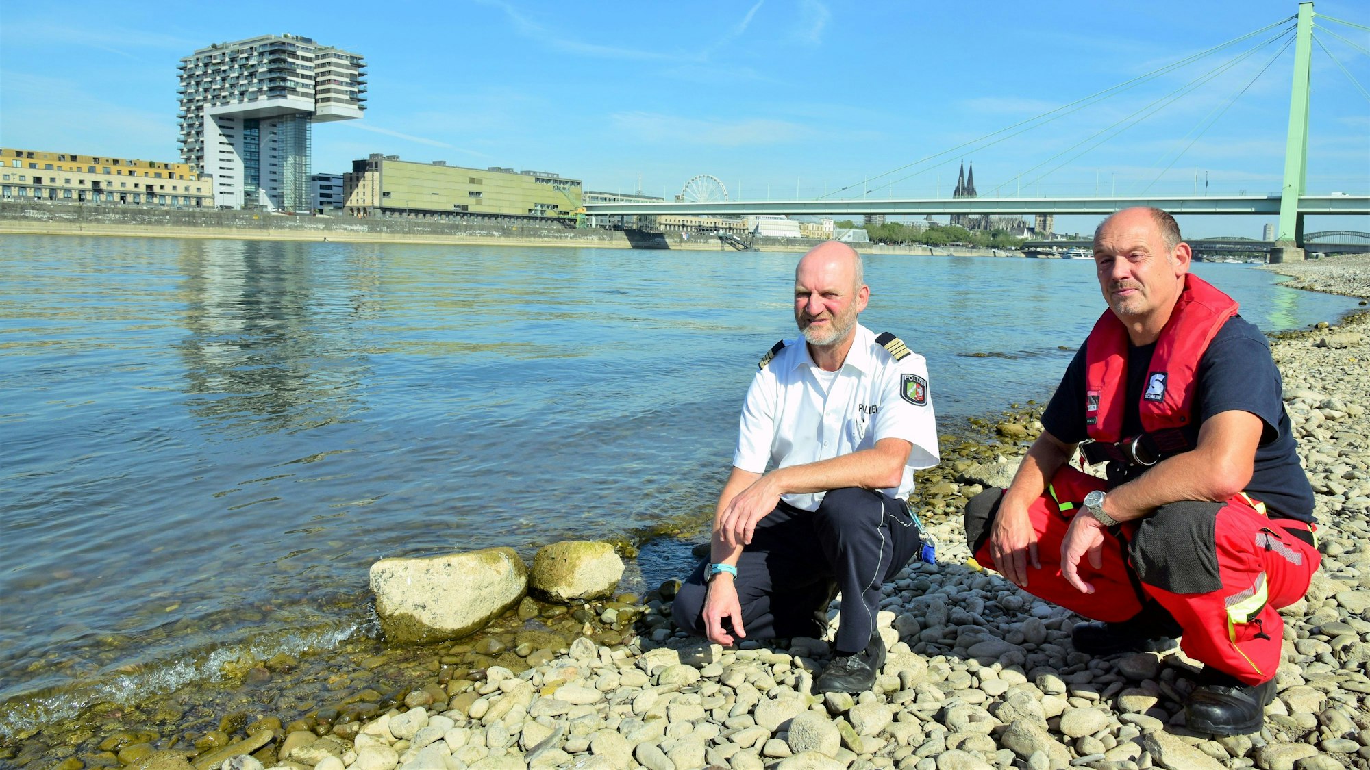 Zwei Kölner Experten hocken direkt am Rheinufer und blicken in die Kamera. Die Gefahren am Rhein sind größer als je zuvor, sagen sie.