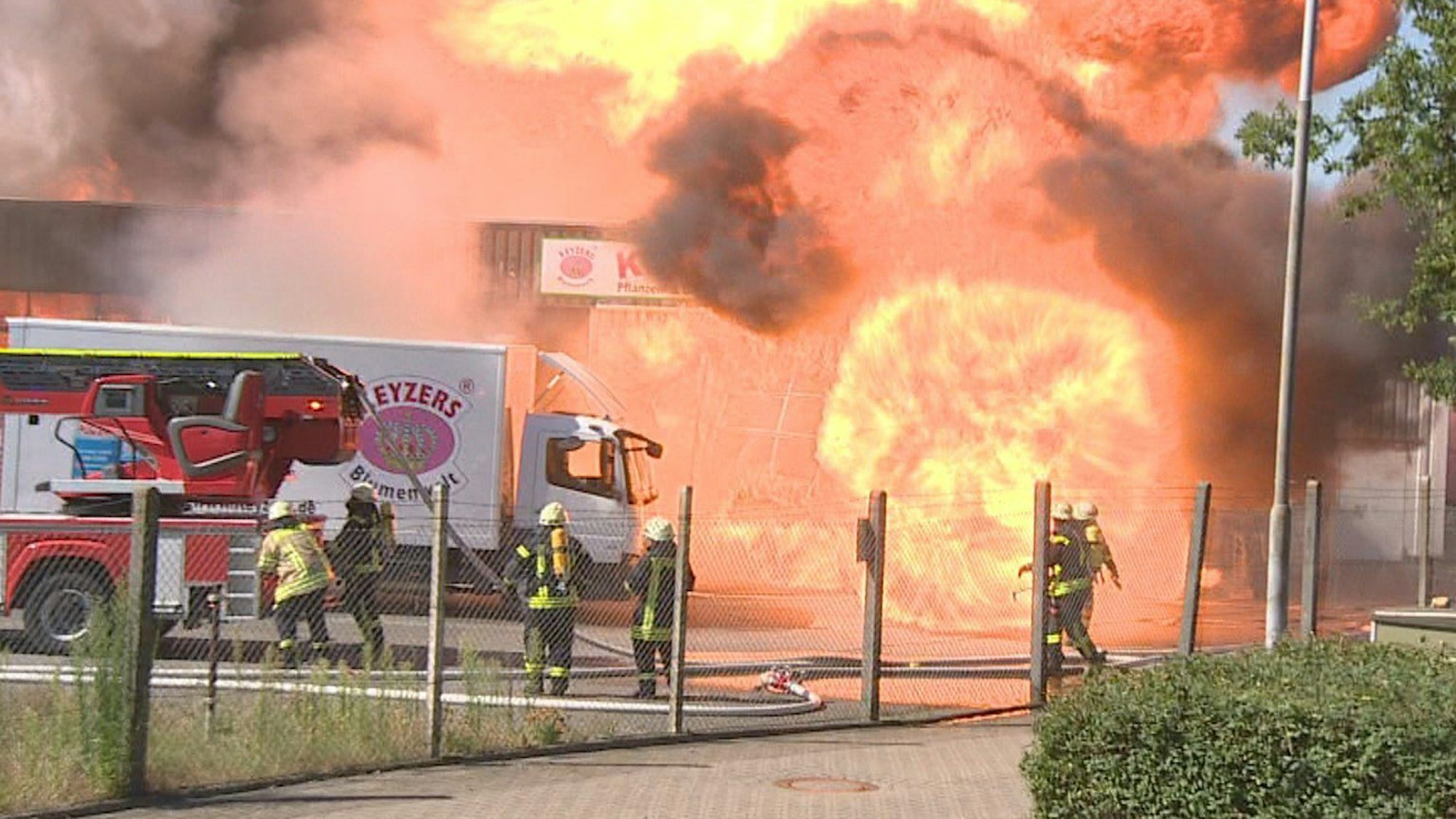 Ein Feuerball schießt aus einen Gebäude, vor dem ein Löschfahrzeug, ein Lkw sowie mehrere Einsatzkräfte der Feuerwehr stehen.