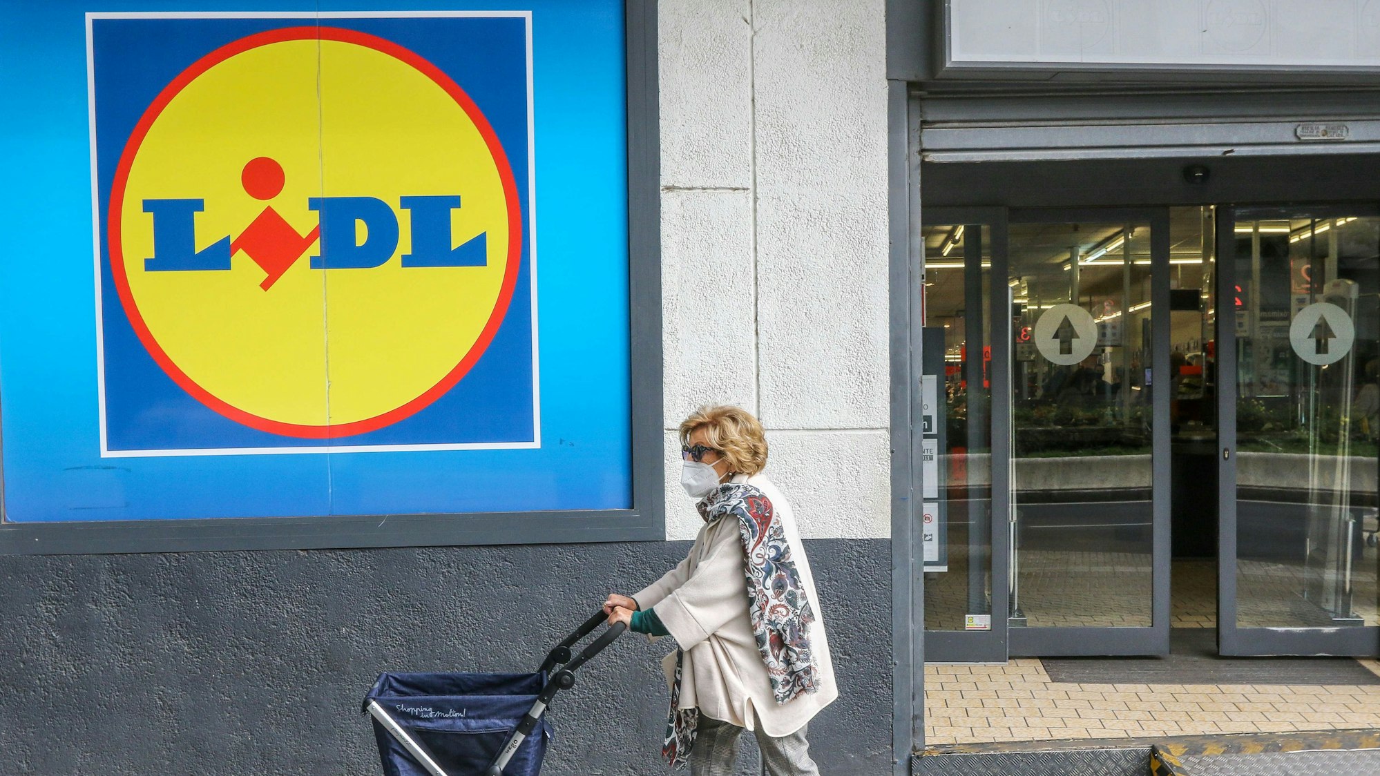 Eine Frau mit Rollator trägt einen Mund-Nasen-Schutz und passiert ein Einkaufsladen von Lidl.
