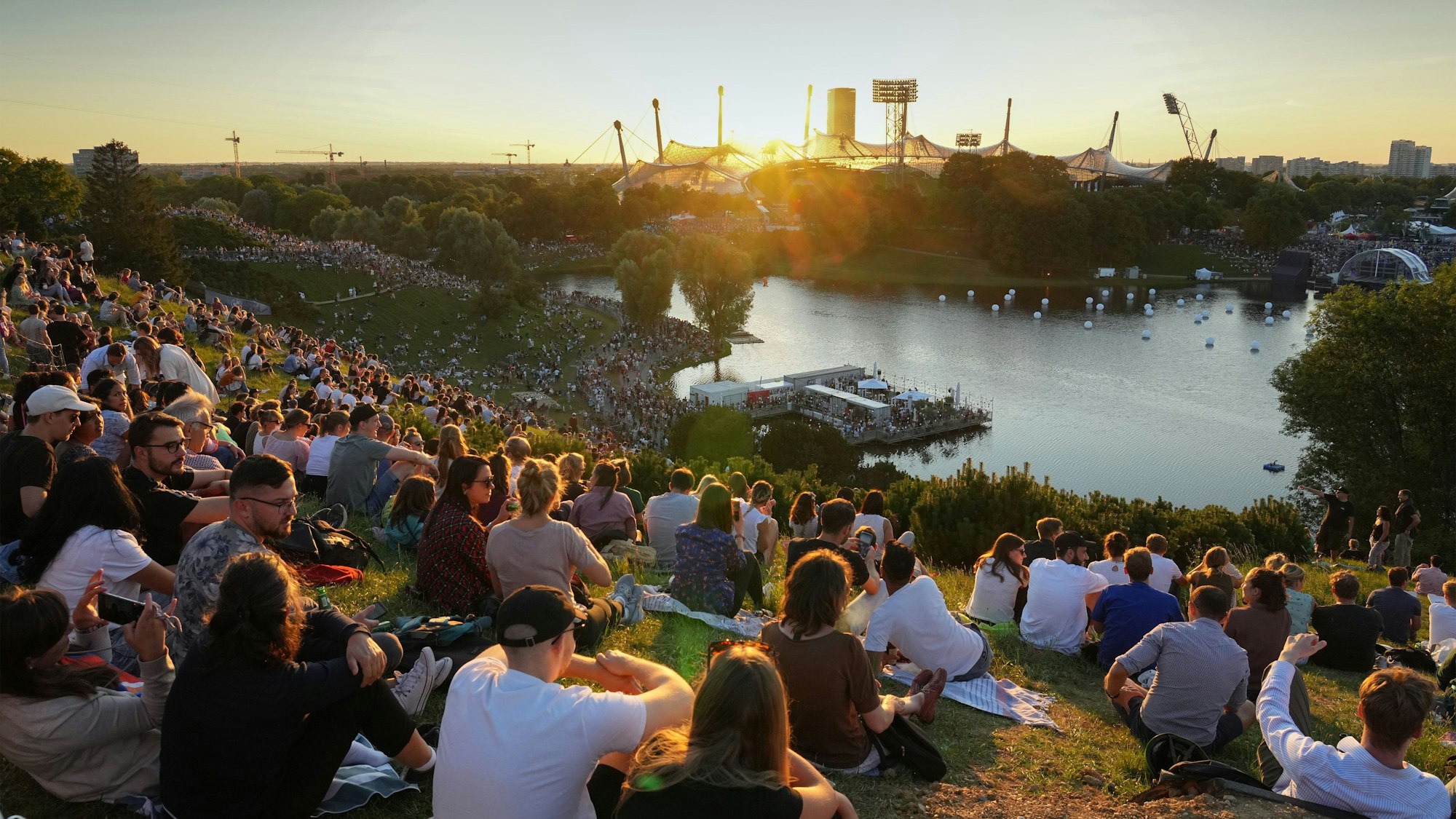 Menschen sitzen auf dem Münchner Olympiaberg und schauen auf den See vor dem Olympiastadion.