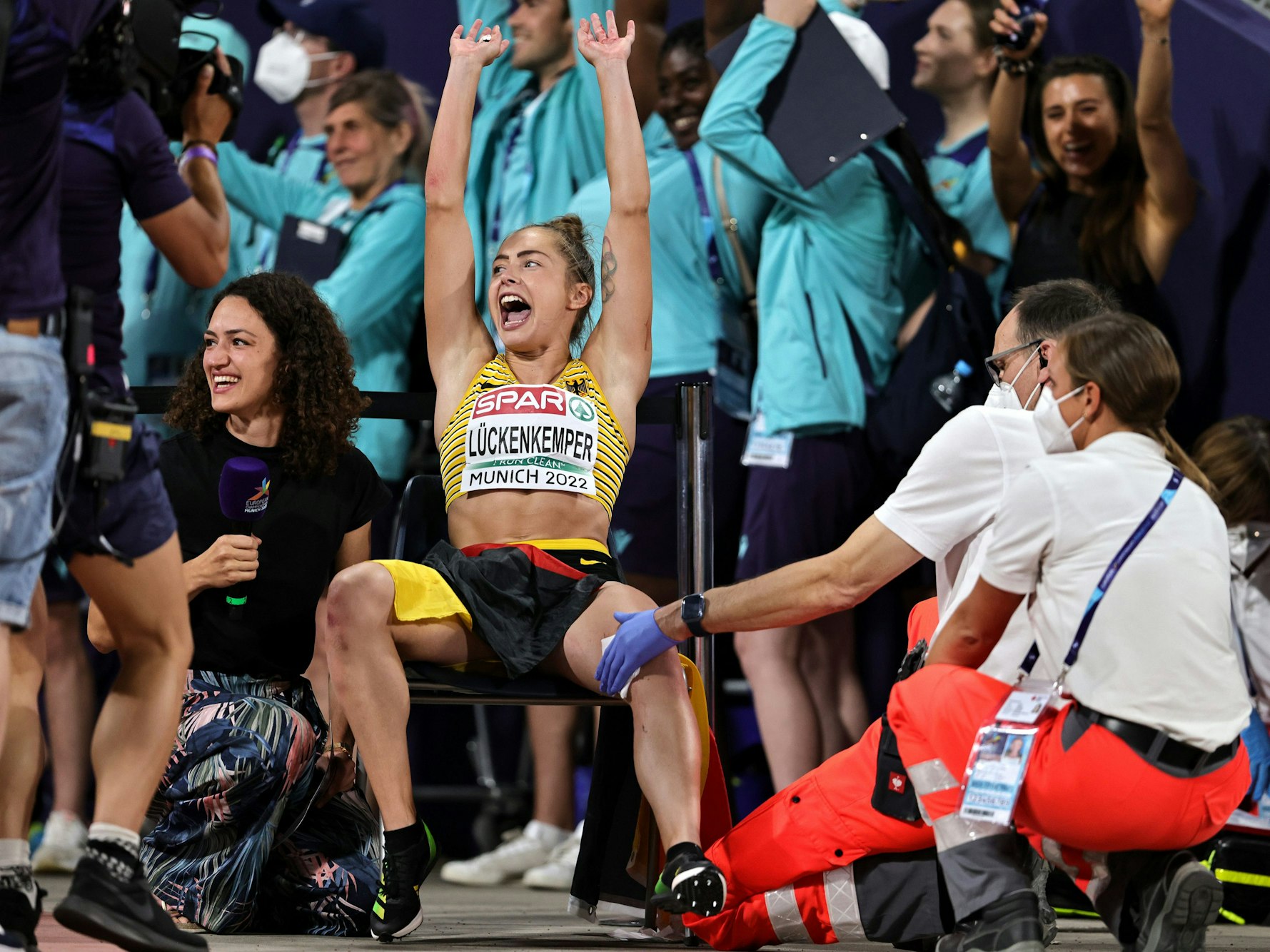 Gina Lückenkemper macht mit den Fans La-Ola. Sanitäter versorgen dabei ihre Wunde.