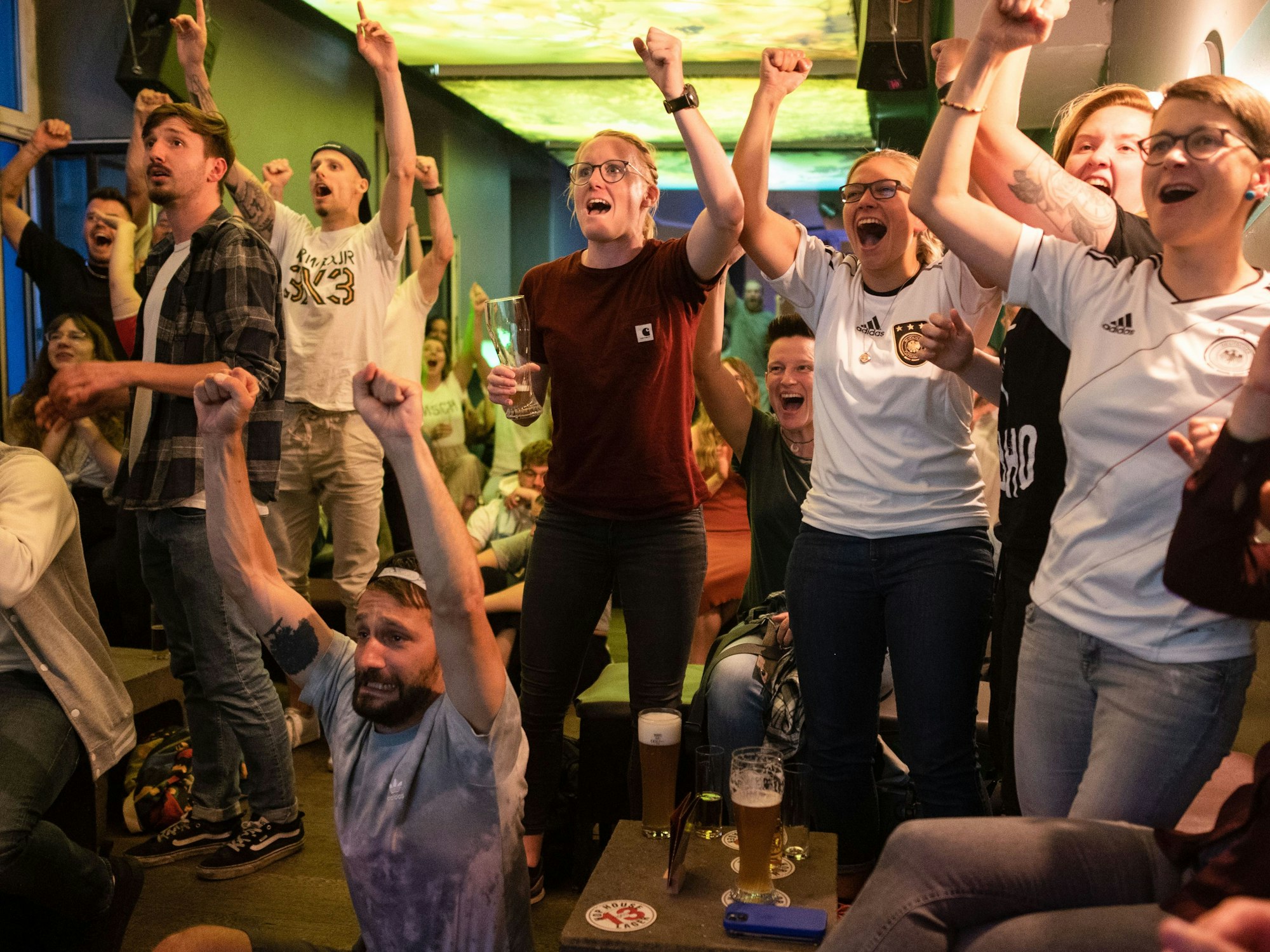 Fußballfans verfolgen ein Fußballspiel in der Kneipe „Gottes grüne Wiese“ und bejubeln einen Tortreffer.