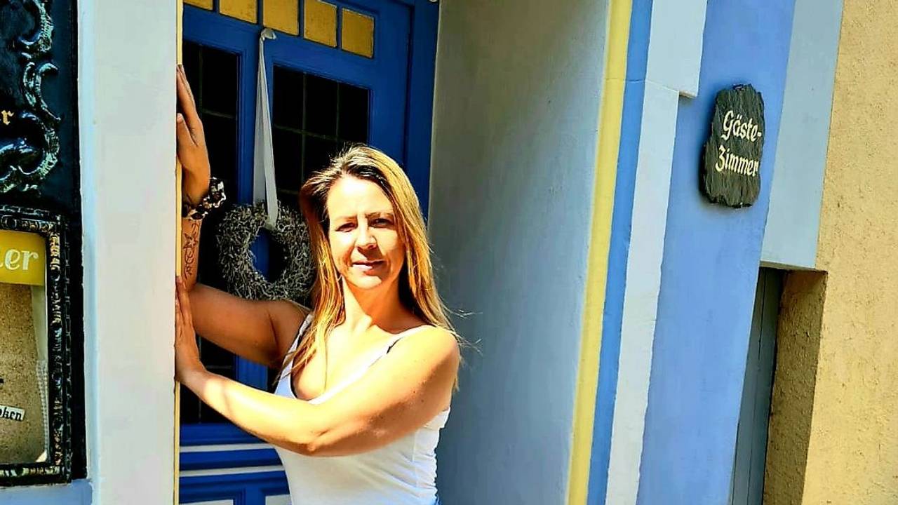 Daniela steht an eine Wand gelehnt vor der Eingangstür eines Hauses.