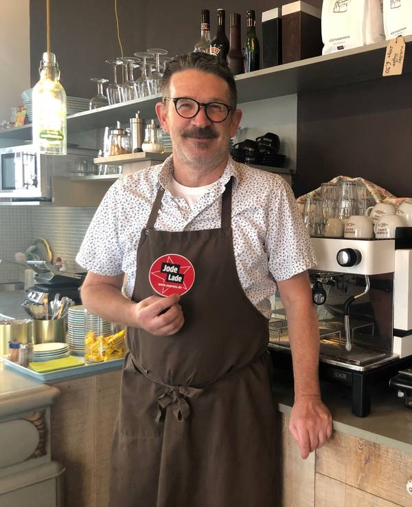 Thomas Kastner steht vor der Kaffeemaschine in seinem Café und hält den „Jode Lade“-Sticker in die Kamera.