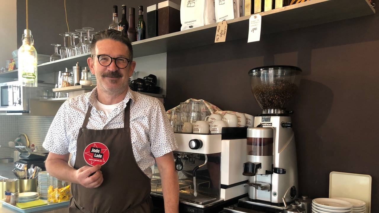 Thomas Kastner steht vor der Kaffeemaschine in seinem Café und hält den „Jode Lade“-Sticker in die Kamera.