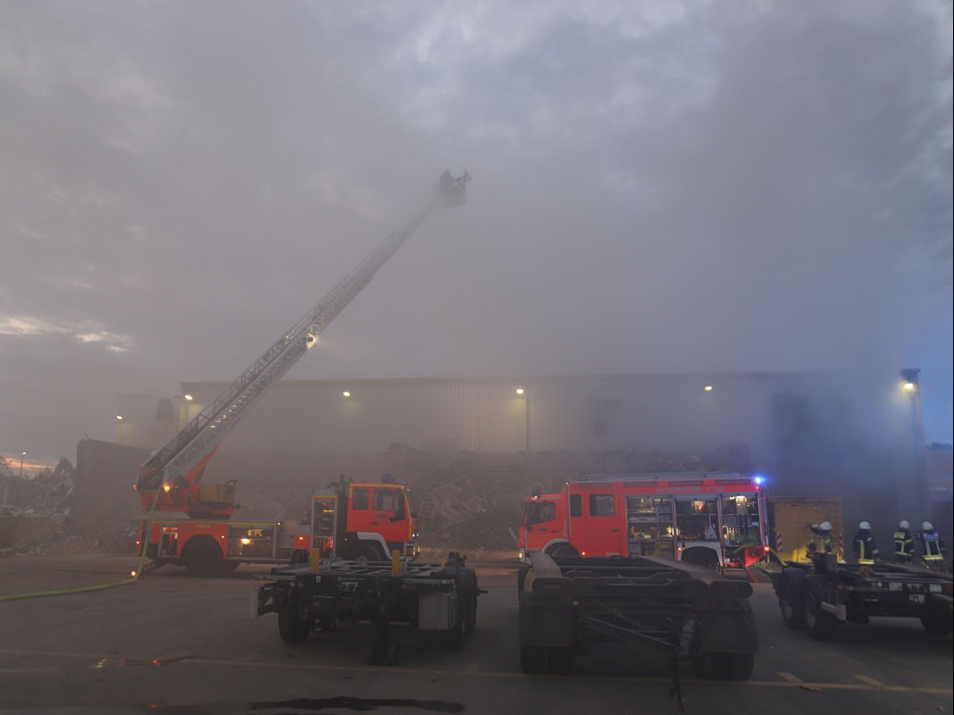 Rauch dringt aus einer Halle, vor der zwei Löschfahrzeuge der Feuerwehr stehen. Es hat die Drehleiter ausgefahren.