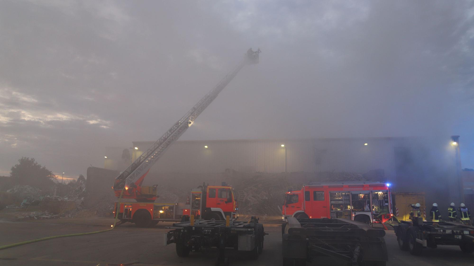 Rauch dringt aus einer Halle, vor der zwei Löschfahrzeuge der Feuerwehr stehen. Es hat die Drehleiter ausgefahren.