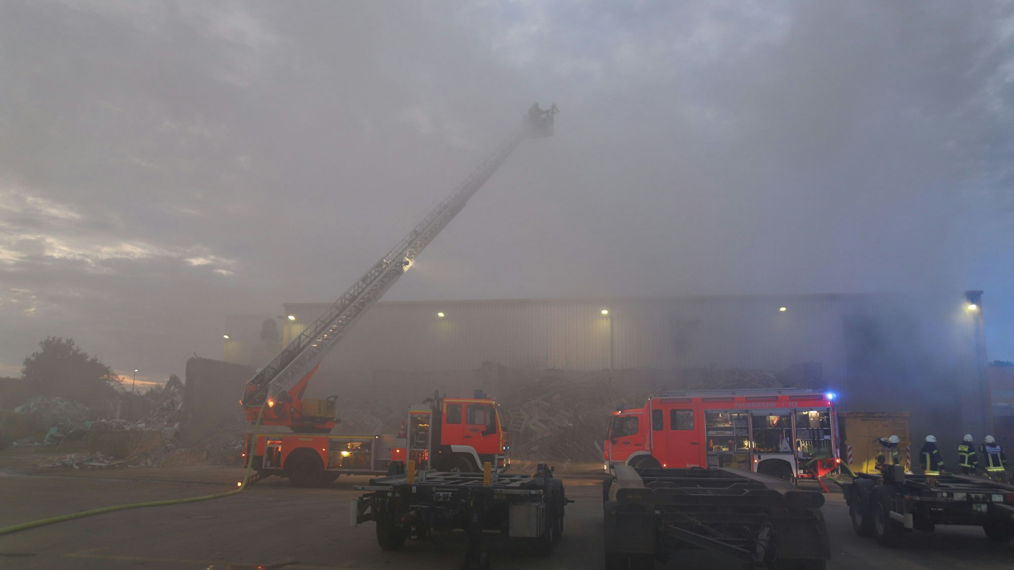 Rauch dringt aus einer Halle, vor der zwei Löschfahrzeuge der Feuerwehr stehen. Es hat die Drehleiter ausgefahren.