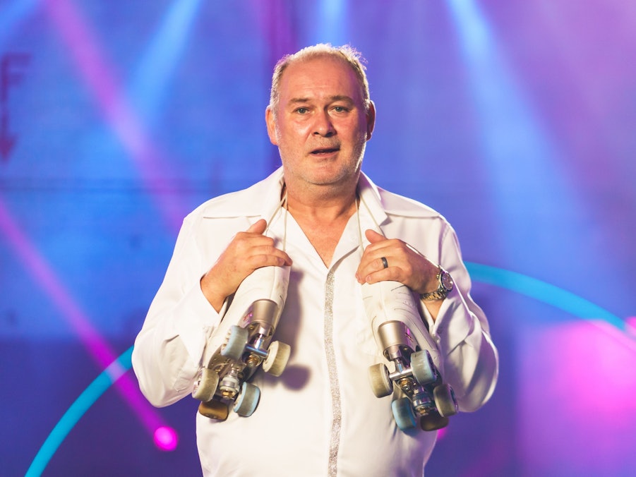 Walter „Waldi“ Lehnertz versucht sich bald bei einer neuen Show im Rollschuhlaufen. Hier trägt er ein weißes Outfit und hat sich seine Rollschuhe um den Nacken gehängt.