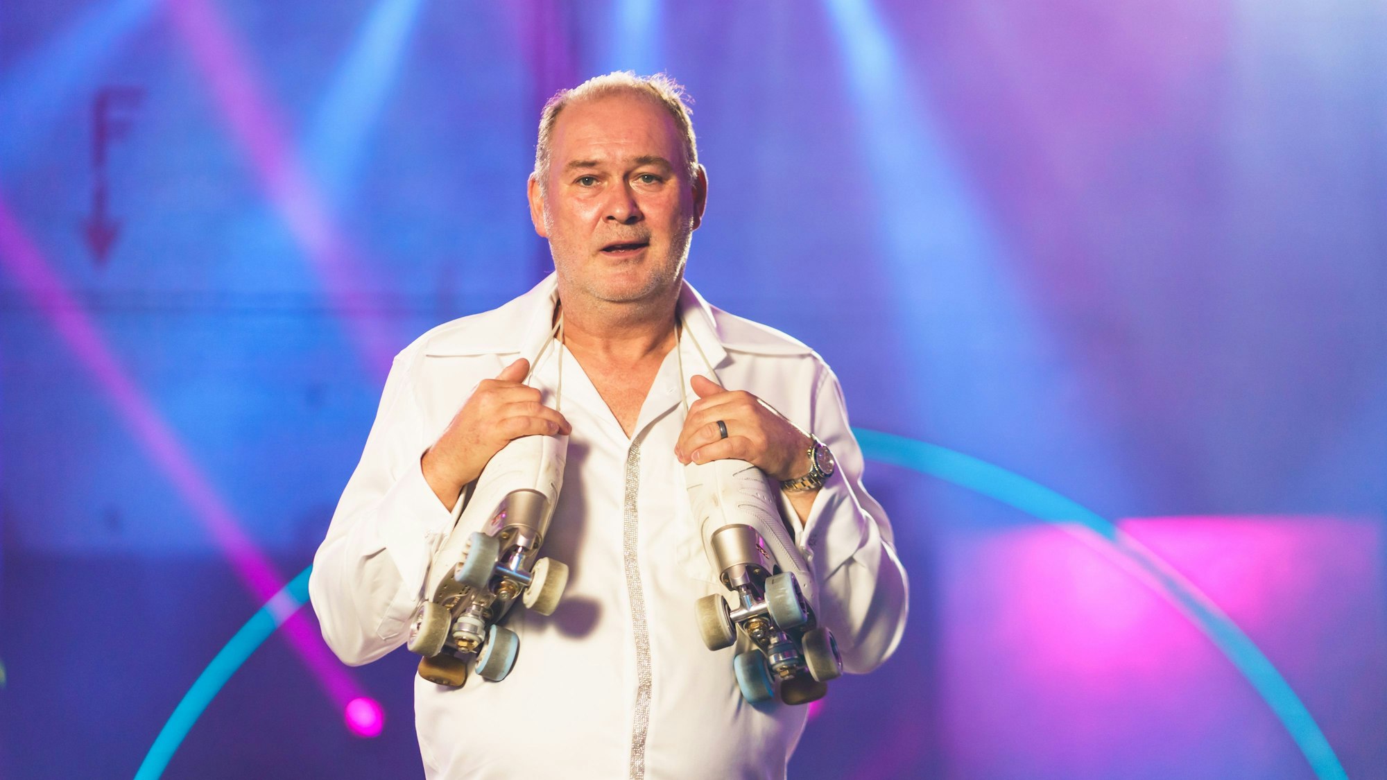 Walter „Waldi“ Lehnertz versucht sich bald bei einer neuen Show im Rollschuhlaufen. Hier trägt er ein weißes Outfit und hat sich seine Rollschuhe um den Nacken gehängt.
