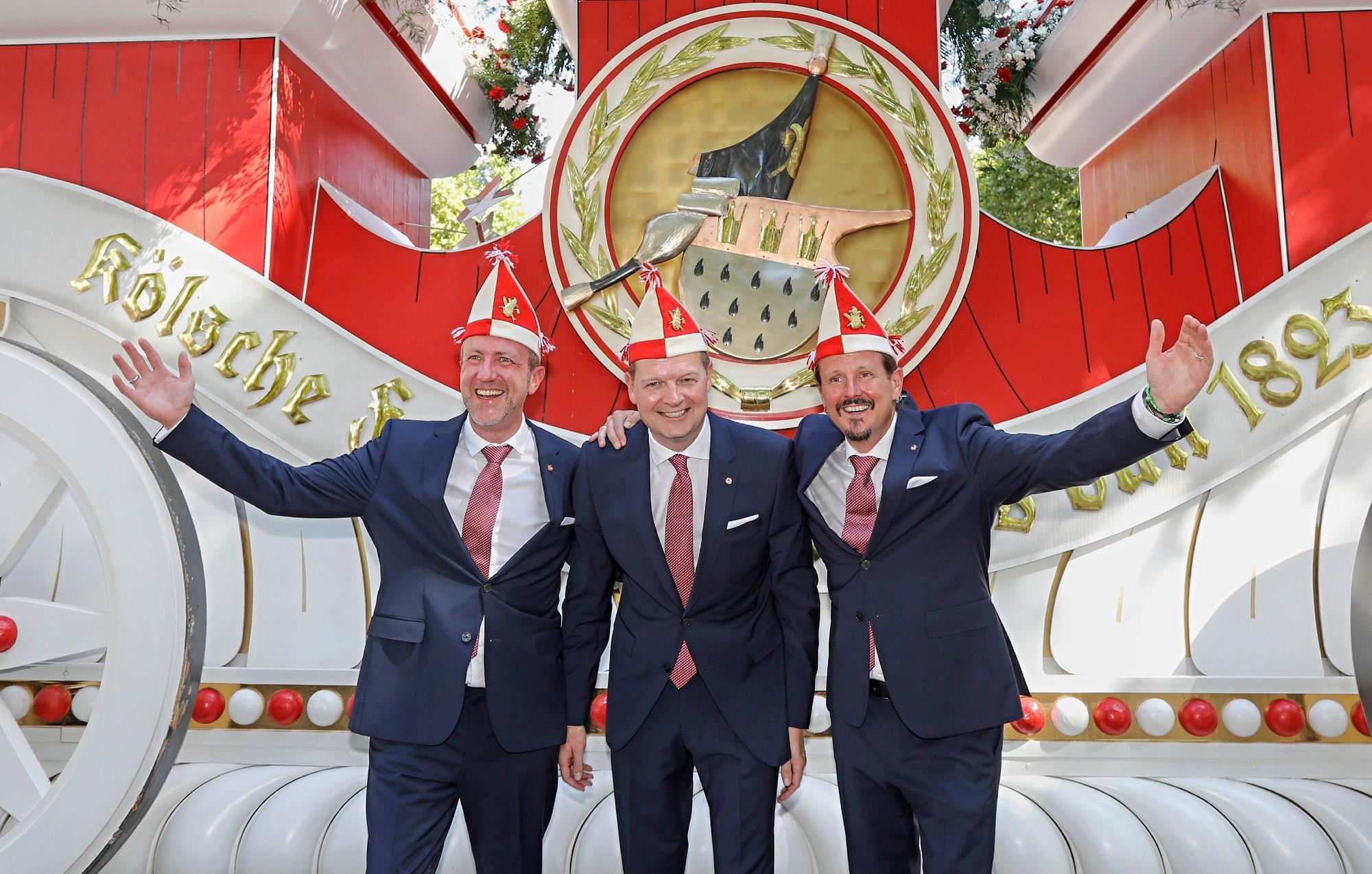 Prinz, Bauer und Jungfrau des neuen Kölner Dreigestirns stehen vor einem Wagen der Roten Funken.