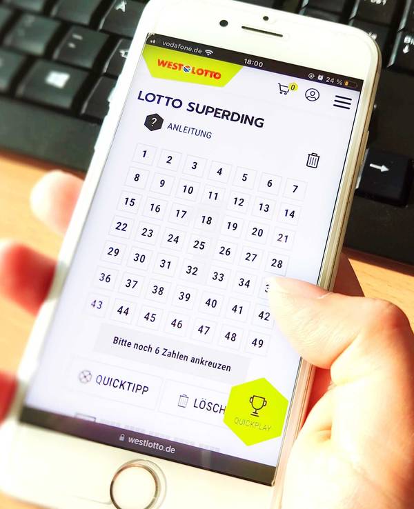 Frauenhand hält Smartphone am Tisch vor einer Tastatur. Bild zum Lotto SuperDing 6aus49 bei WestLotto Artikel .
