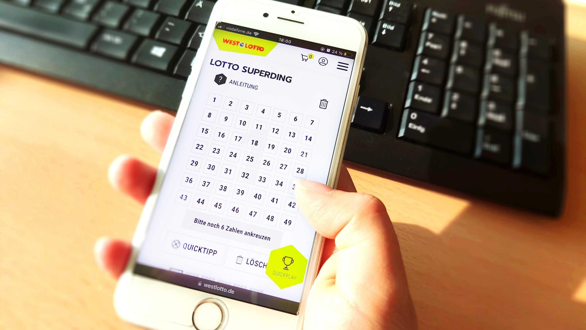 Frauenhand hält Smartphone am Tisch vor einer Tastatur. Bild zum Lotto SuperDing 6aus49 bei WestLotto Artikel .