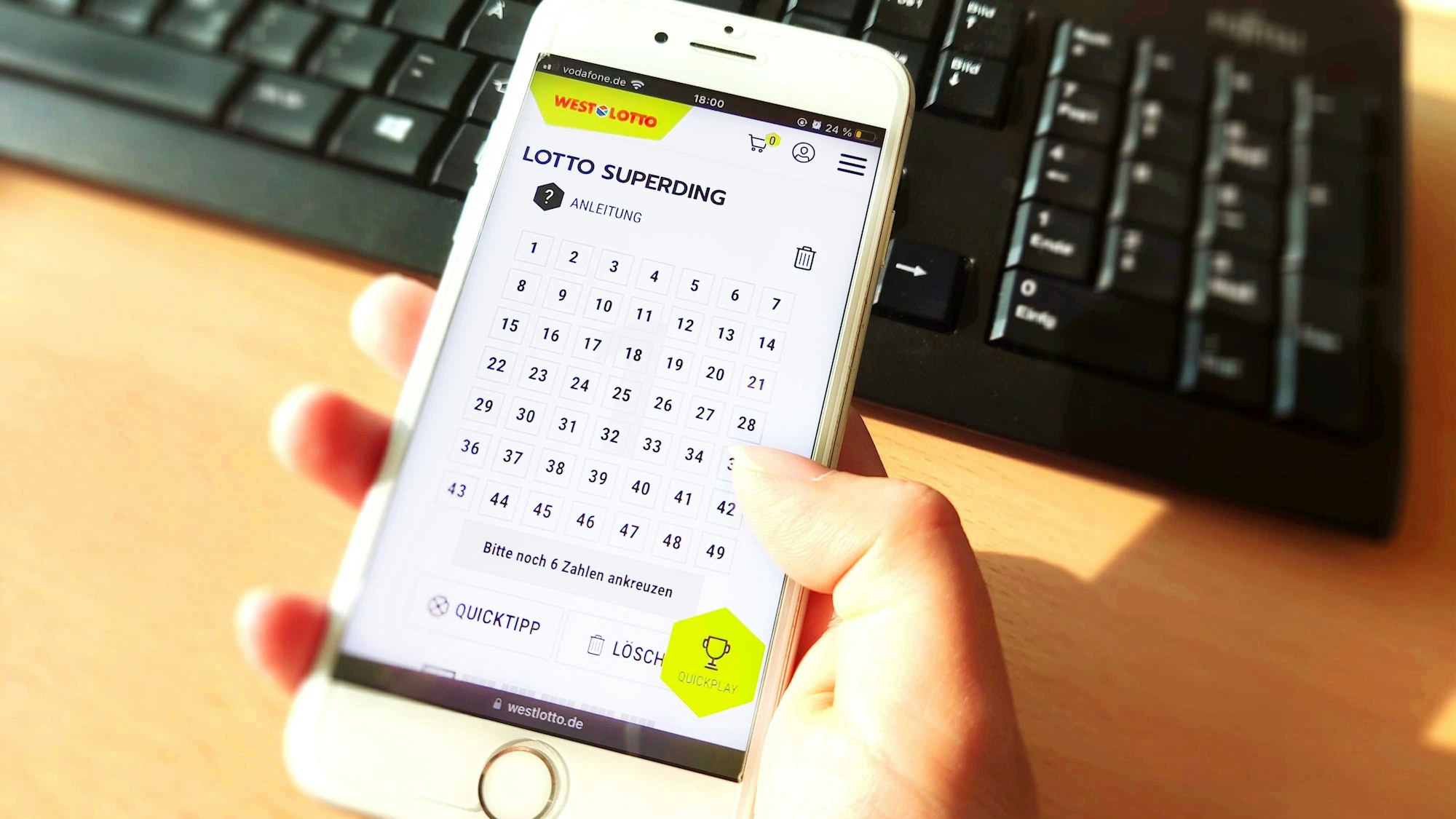 Frauenhand hält Smartphone am Tisch vor einer Tastatur. Bild zum Lotto SuperDing 6aus49 bei WestLotto Artikel .
