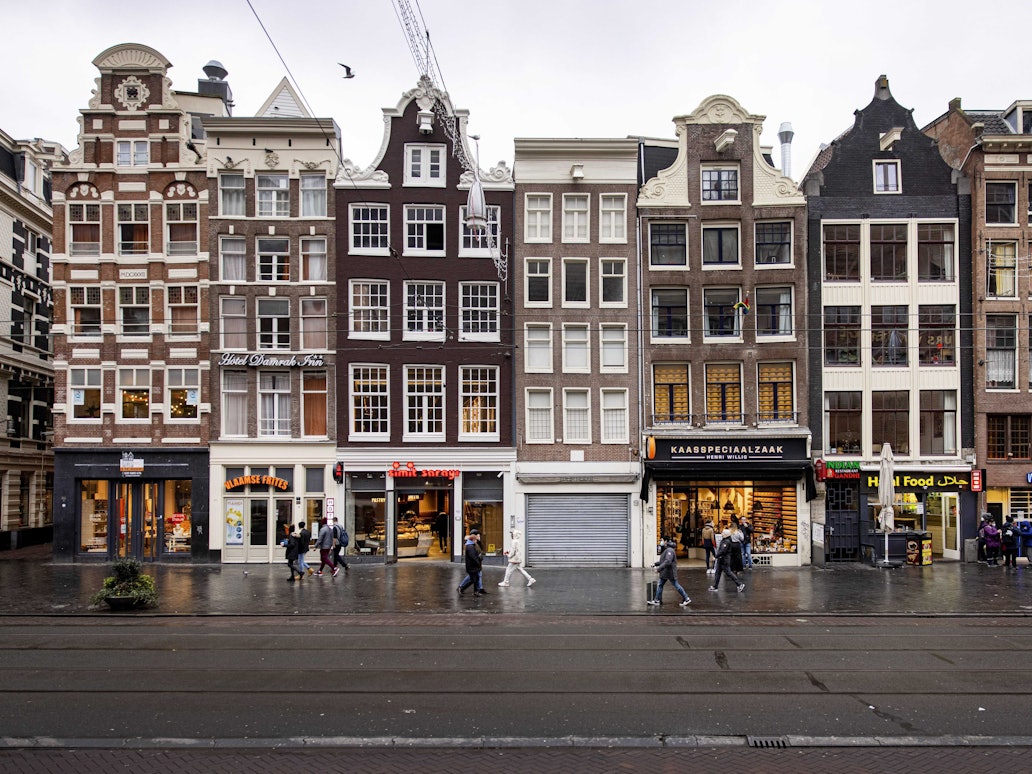 Das Bild zeigt eine Häuserreihe in den Niederlanden mit Ladenlokalen im Erdgeschoss. Auf dem Bürgersteig laufen Passanten und Passantinnen.