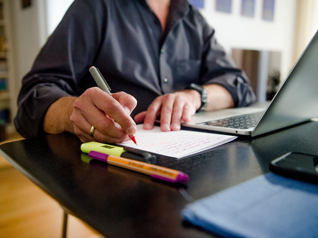 Ein Mann sitzt zuhause an einem Esstisch. Er schreibt mit einem Stift auf ein Blatt Papier neben einem Laptop