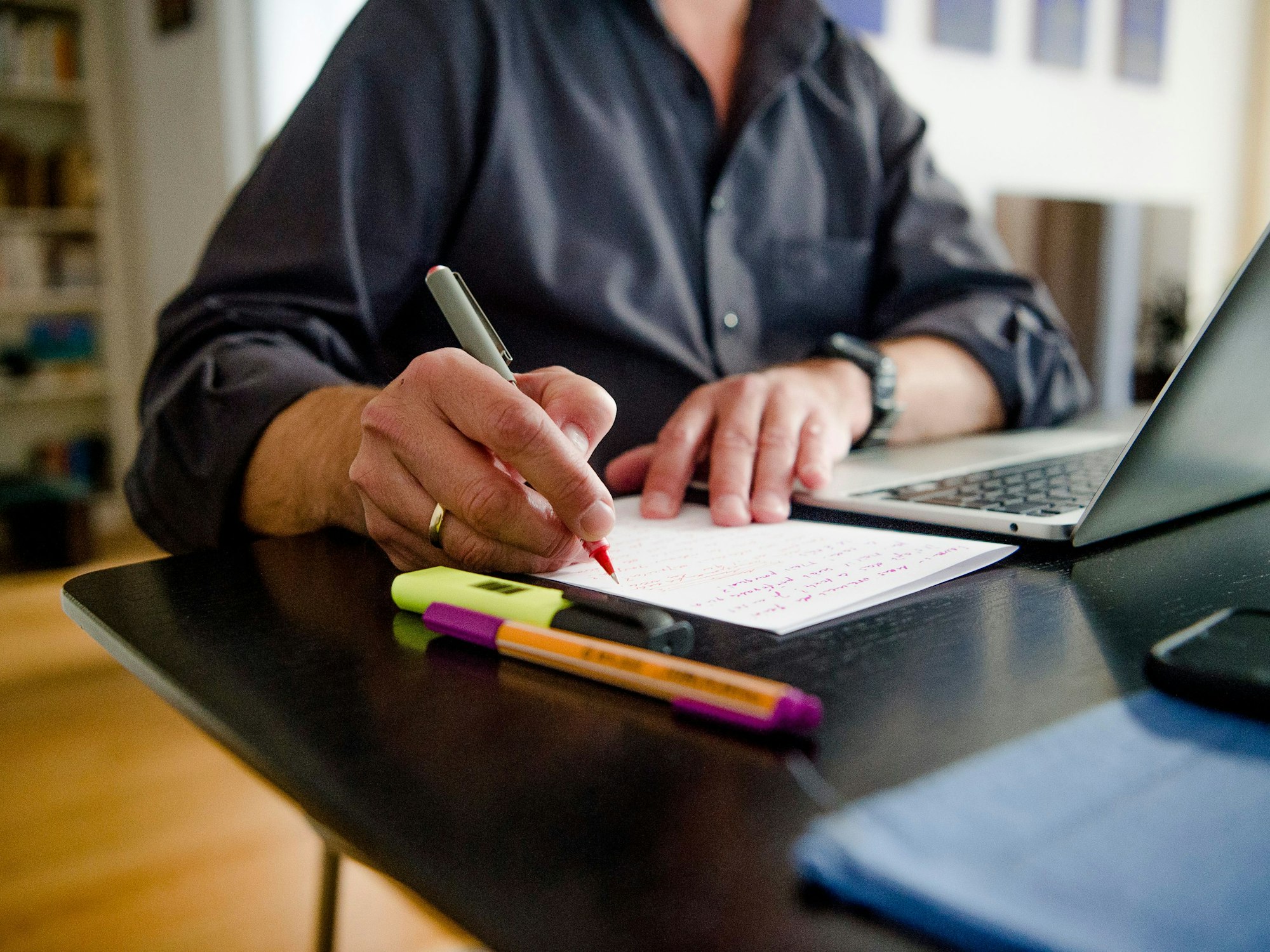 Ein Mann sitzt zuhause an einem Esstisch. Er schreibt mit einem Stift auf ein Blatt Papier neben einem Laptop