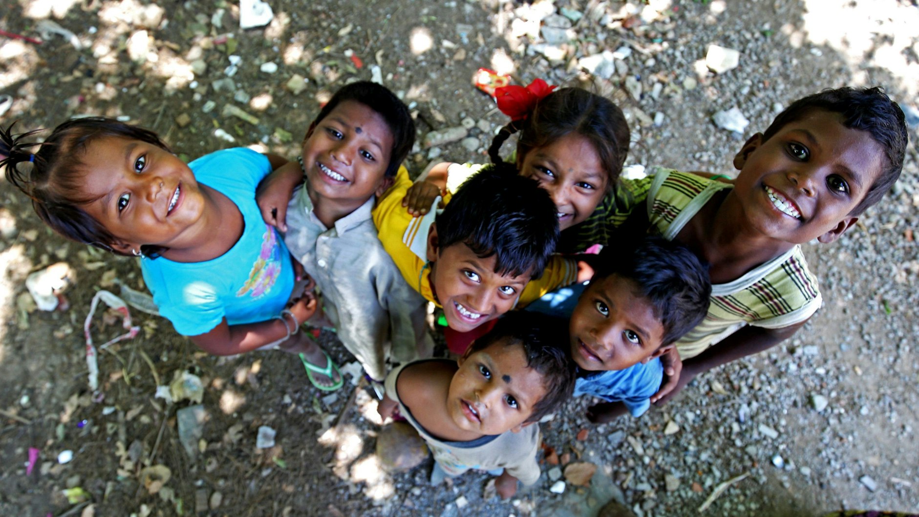 Unser Symbolfoto vom 24. Mai 2016 zeigt sieben indische Kinder, die in die Kamera blicken. Manche lächeln, andere schauen ängstlich. Sie stehen auf einem sandigen Untergrund mit Scherben, Steinen und Müll.