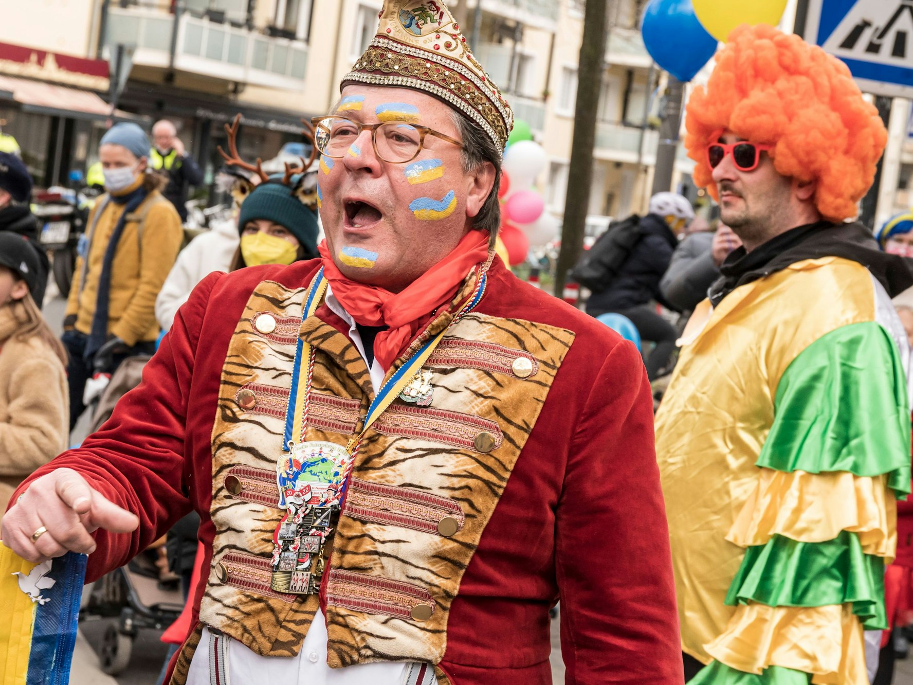 Hans Mörtter verkleidet und mit aufgemalten Ukraine-Flaggen in Gesicht auf der Straße, umgeben von verkleideten Menschen.