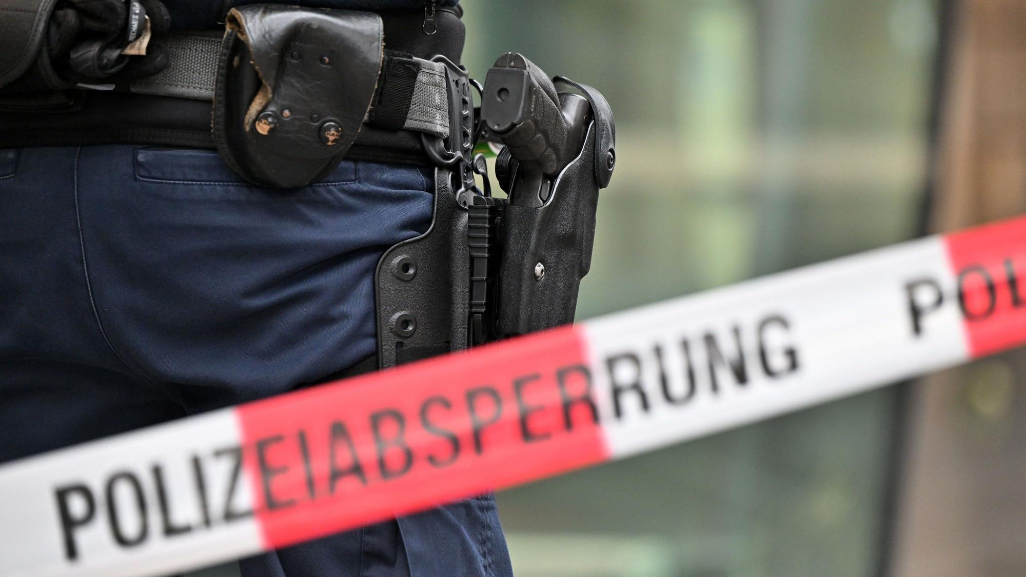 Das undatierte Symbolfoto zeigt ein rot-weißes Flatterband mit der Aufschrift „Polizeiabsperrung“. Dahinter steht eine Person mit einer Waffe im Holster.