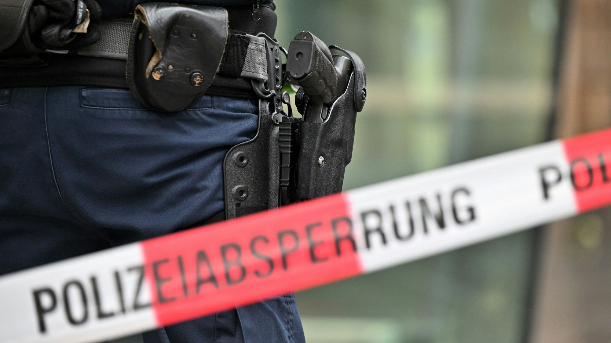Das undatierte Symbolfoto zeigt ein rot-weißes Flatterband mit der Aufschrift „Polizeiabsperrung“. Dahinter steht eine Person mit einer Waffe im Holster.