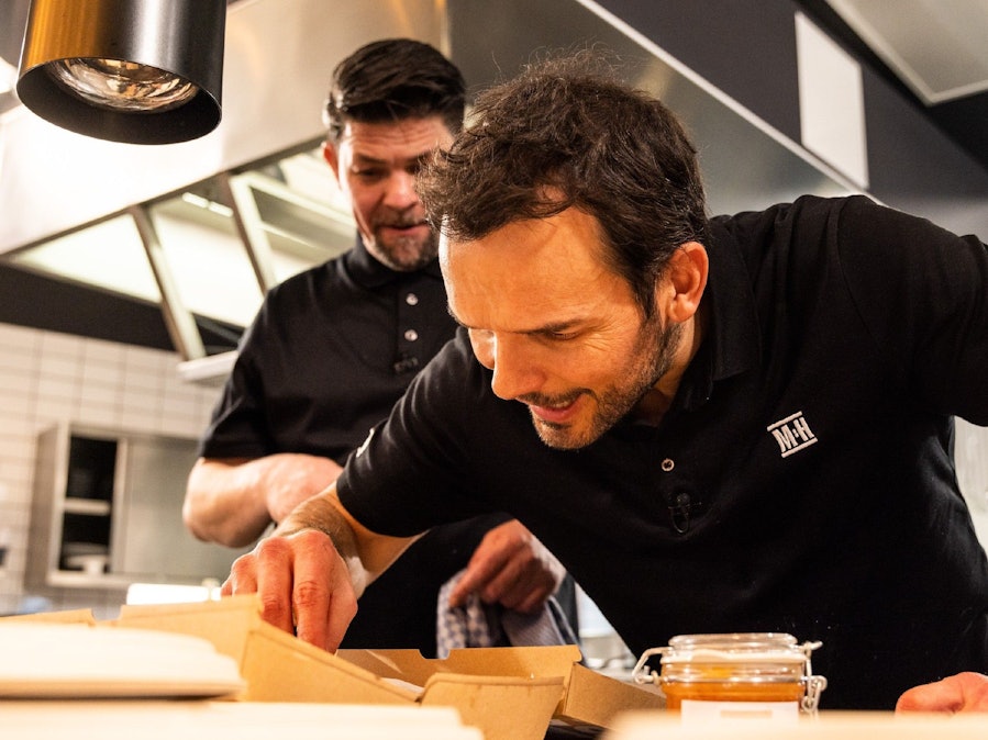 Tim Mälzer und Steffen Henssler stehen in der Kochshow „Mälzer und Henssler liefern ab“ vor einem Tisch und begutachten Zutaten.