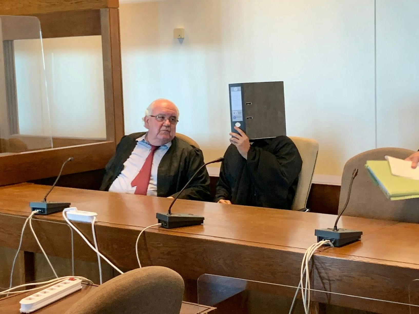 Der angeklagte Anil G. versteckt sich zwischen seinen Anwälten hinter einem Aktenordner.