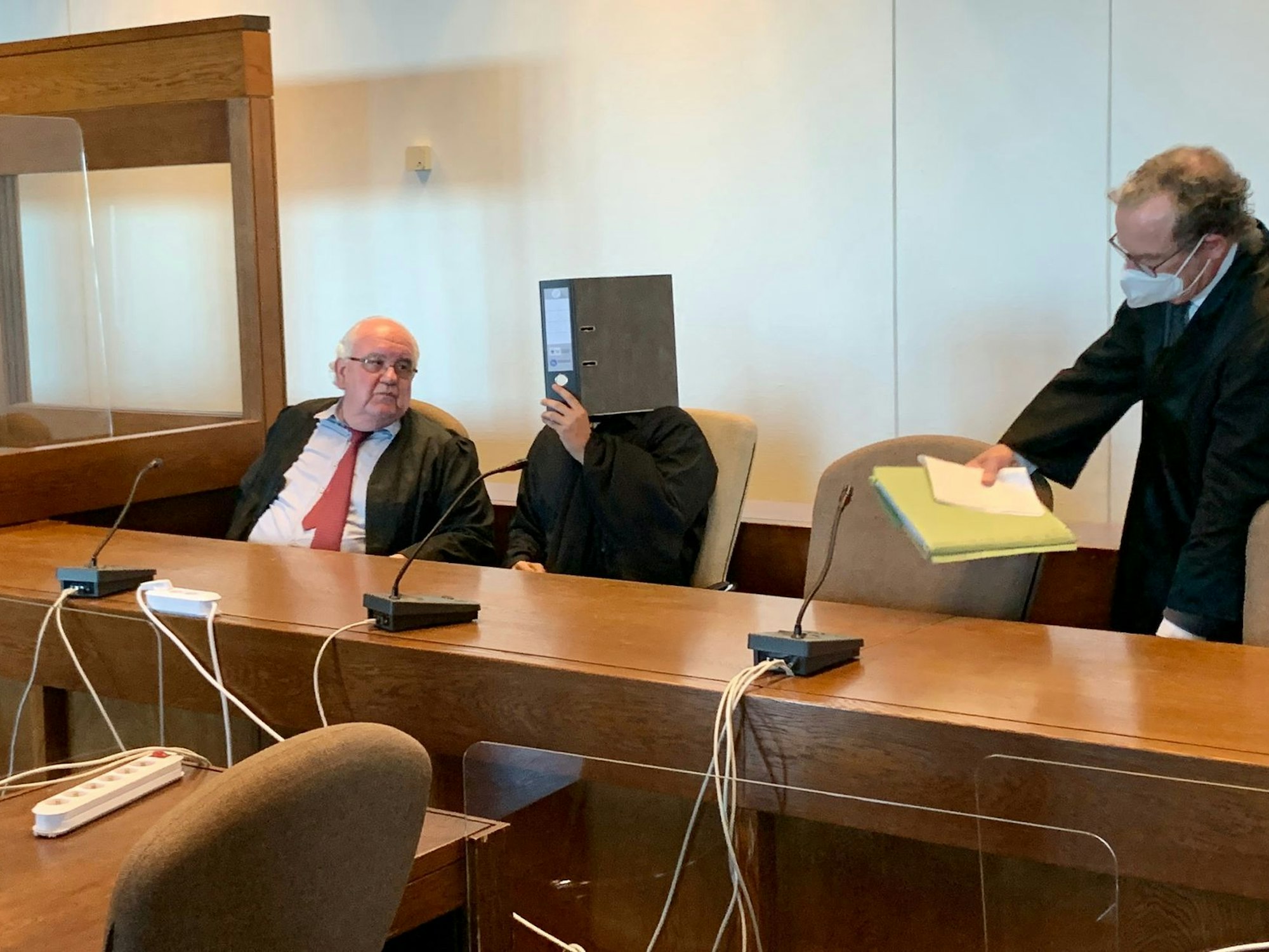 Der angeklagte Anil G. versteckt sich zwischen seinen Anwälten hinter einem Aktenordner.