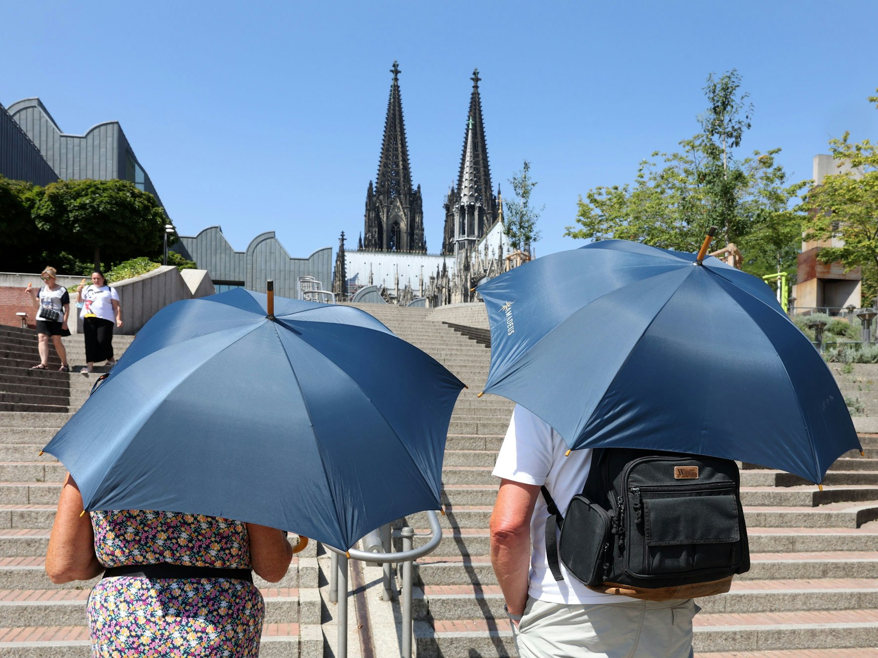 Zwei Touristen laufen mit Sonnenschirmen in Richtung Kölner Dom.