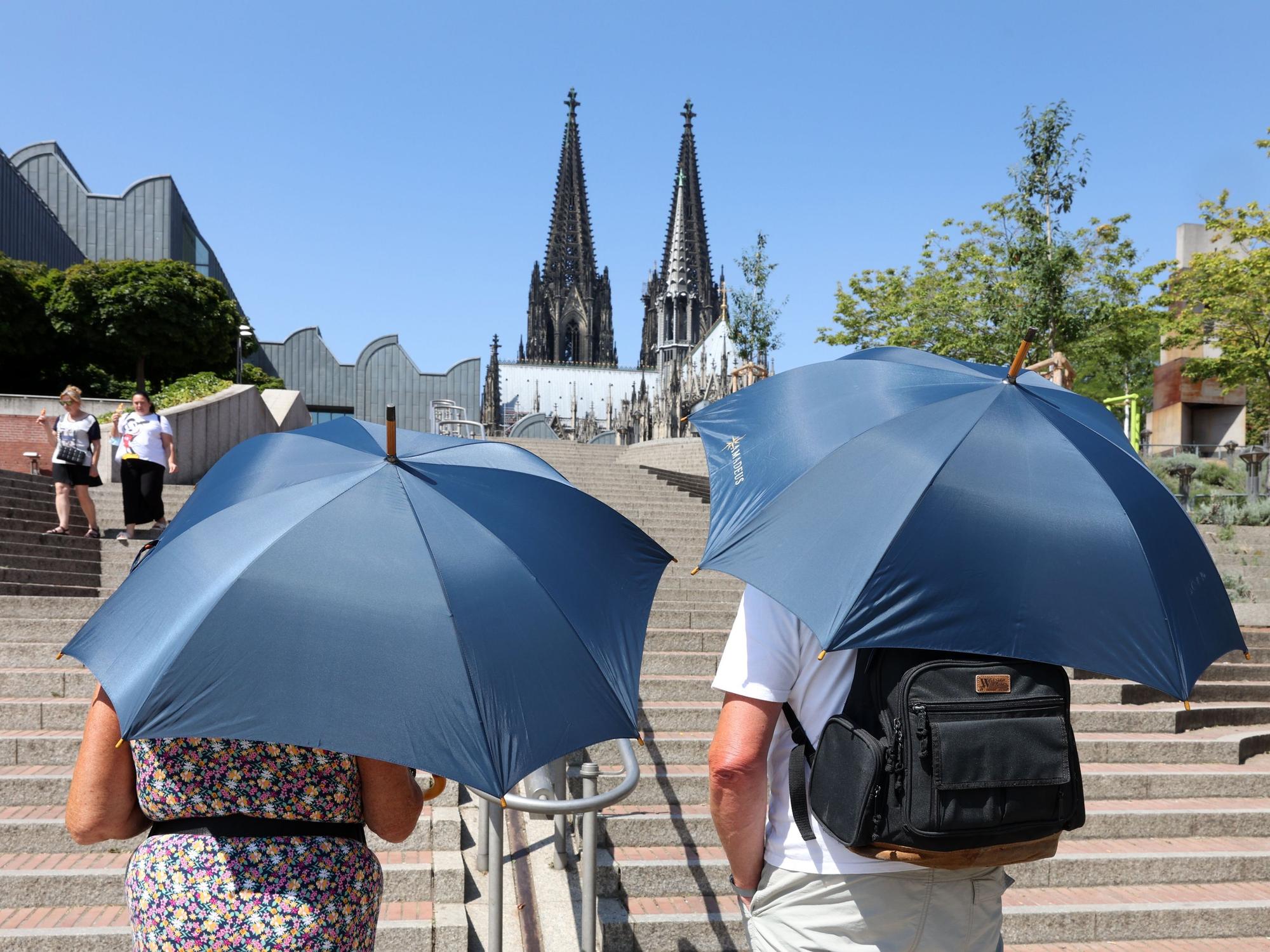Zwei Touristen laufen mit Sonnenschirmen in Richtung Kölner Dom.