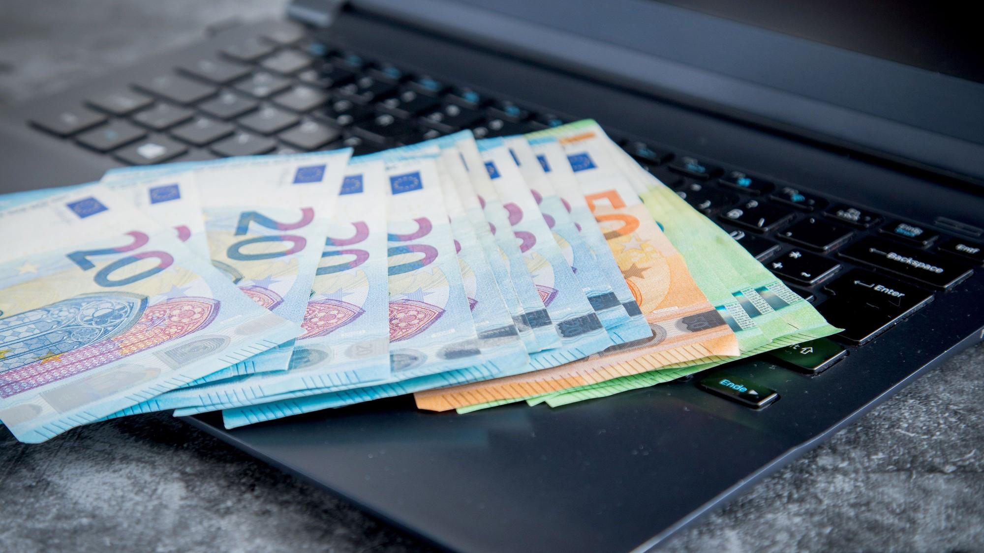 Das Symbolfoto von 2021 zeigt einen schwarzen aufgeklappten Laptop, auf dem mehrere Geldscheine liegen.