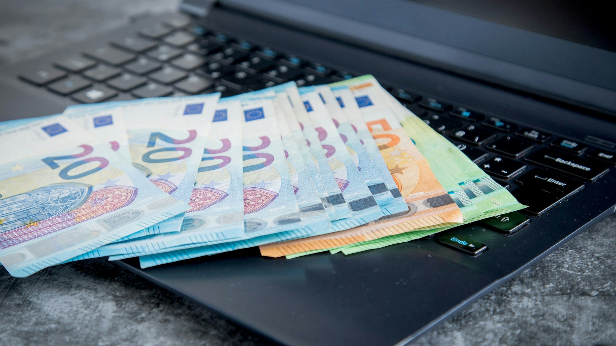 Das Symbolfoto von 2021 zeigt einen schwarzen aufgeklappten Laptop, auf dem mehrere Geldscheine liegen.