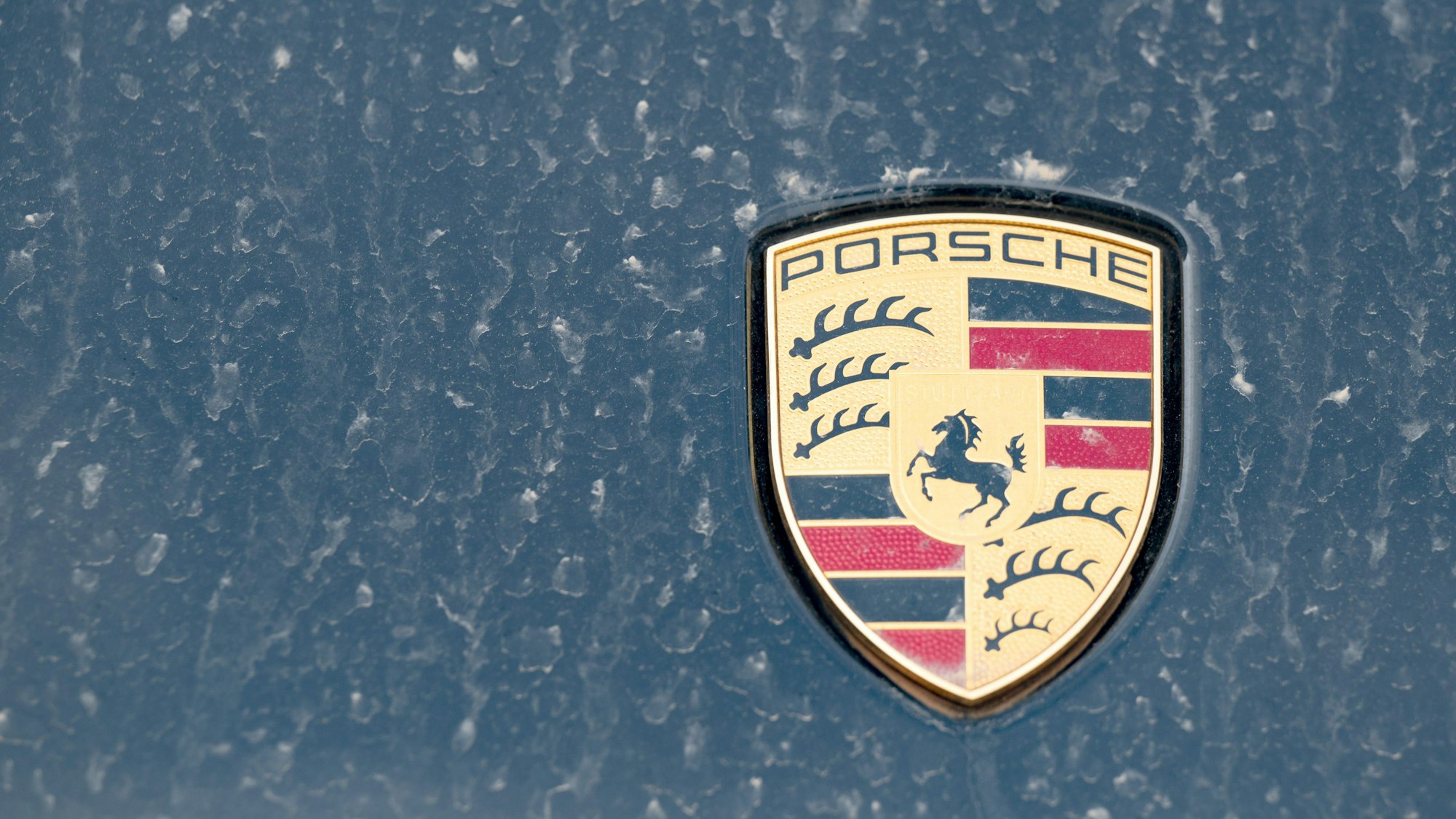Ein Logo des Fahrzeugbauers Porsche ist an einer mit Wüstenstaub bedeckten Motohaube eines Sportwagens angebracht.