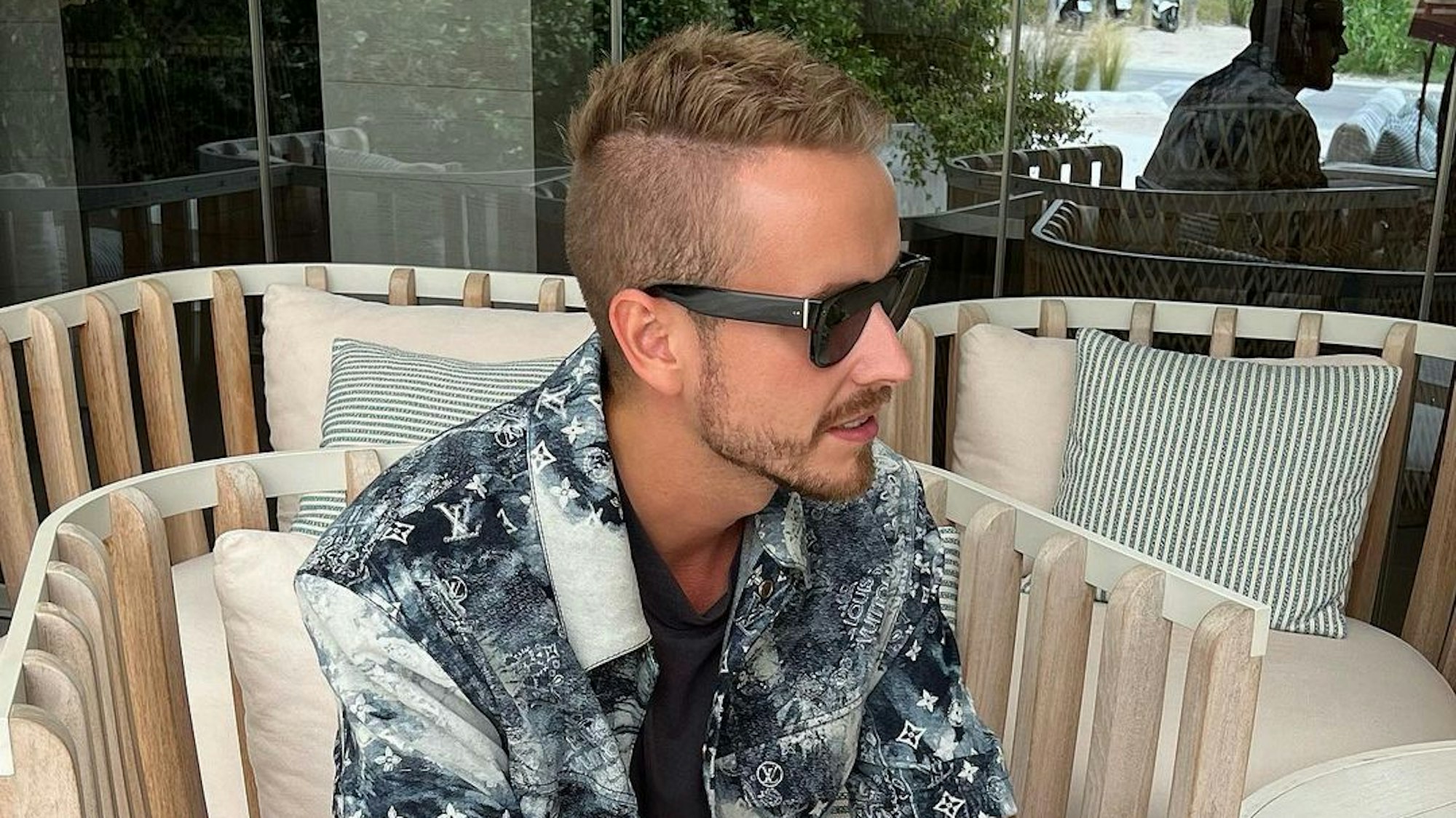 Influencer Julian Claßen, hier auf einem Instagram-Bild von Juni 2022, macht mit seiner neuen Freundin Tanja Makaric den nächsten Schritt. Julian sitzt in einem Korbstuhl, er trägt ein schwarz-weiß gemustertes Hemd und eine schwarze Sonnenbrille.
