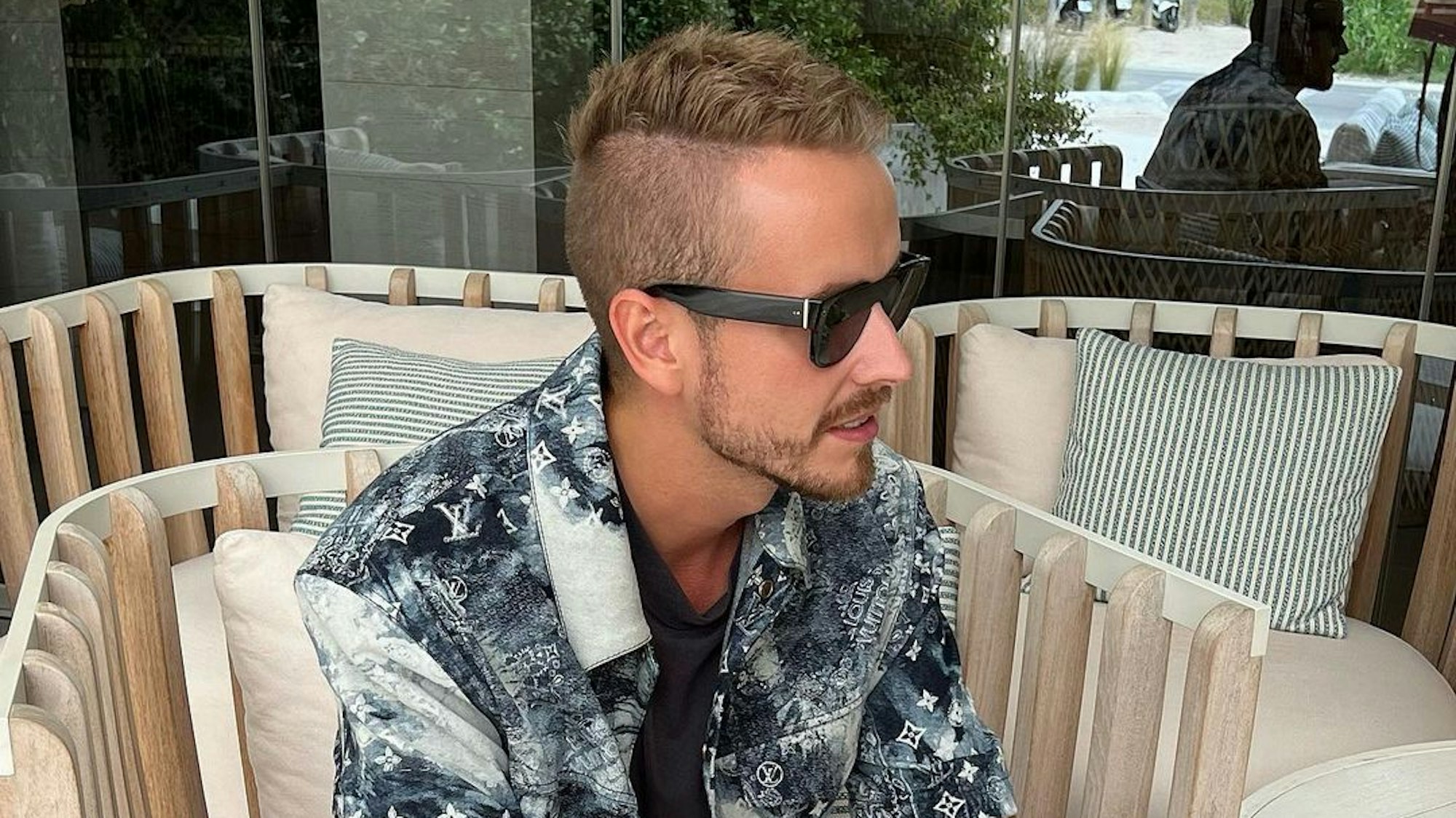 Influencer Julian Claßen, hier auf einem Instagram-Bild von Juni 2022, macht mit seiner neuen Freundin Tanja Makaric den nächsten Schritt. Julian sitzt in einem Korbstuhl, er trägt ein schwarz-weiß gemustertes Hemd und eine schwarze Sonnenbrille.