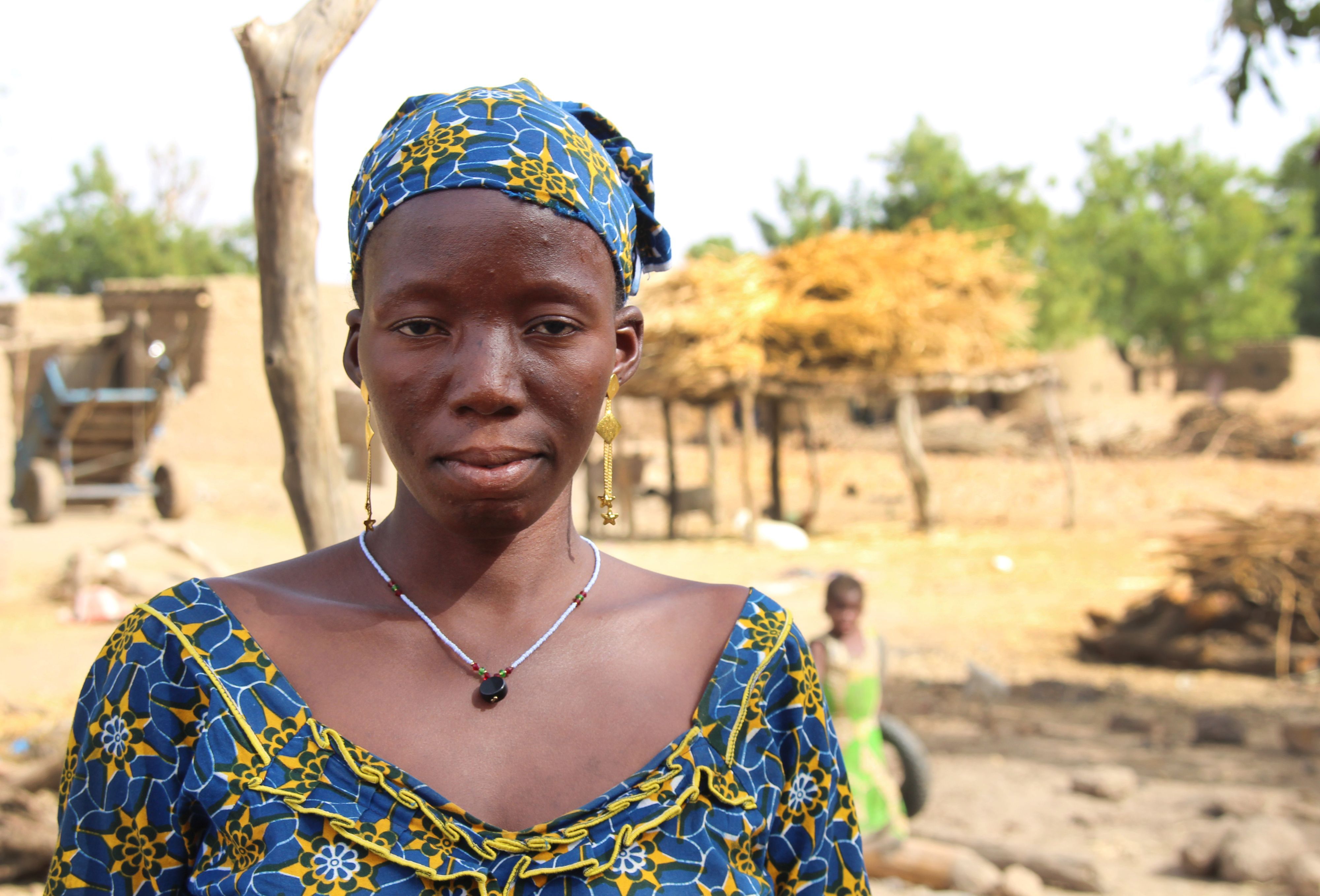 Das Bild zeigt eine junge Afrikanerin in traditioneller Kleidung vor dem Hintergrund eines Dorfes. Es handelt sich um Sanamba aus Mali, hier aufgenommen am 20. März 2018. Sie wurde schon im Alter von 14 Jahren zwangsverheiratet.
