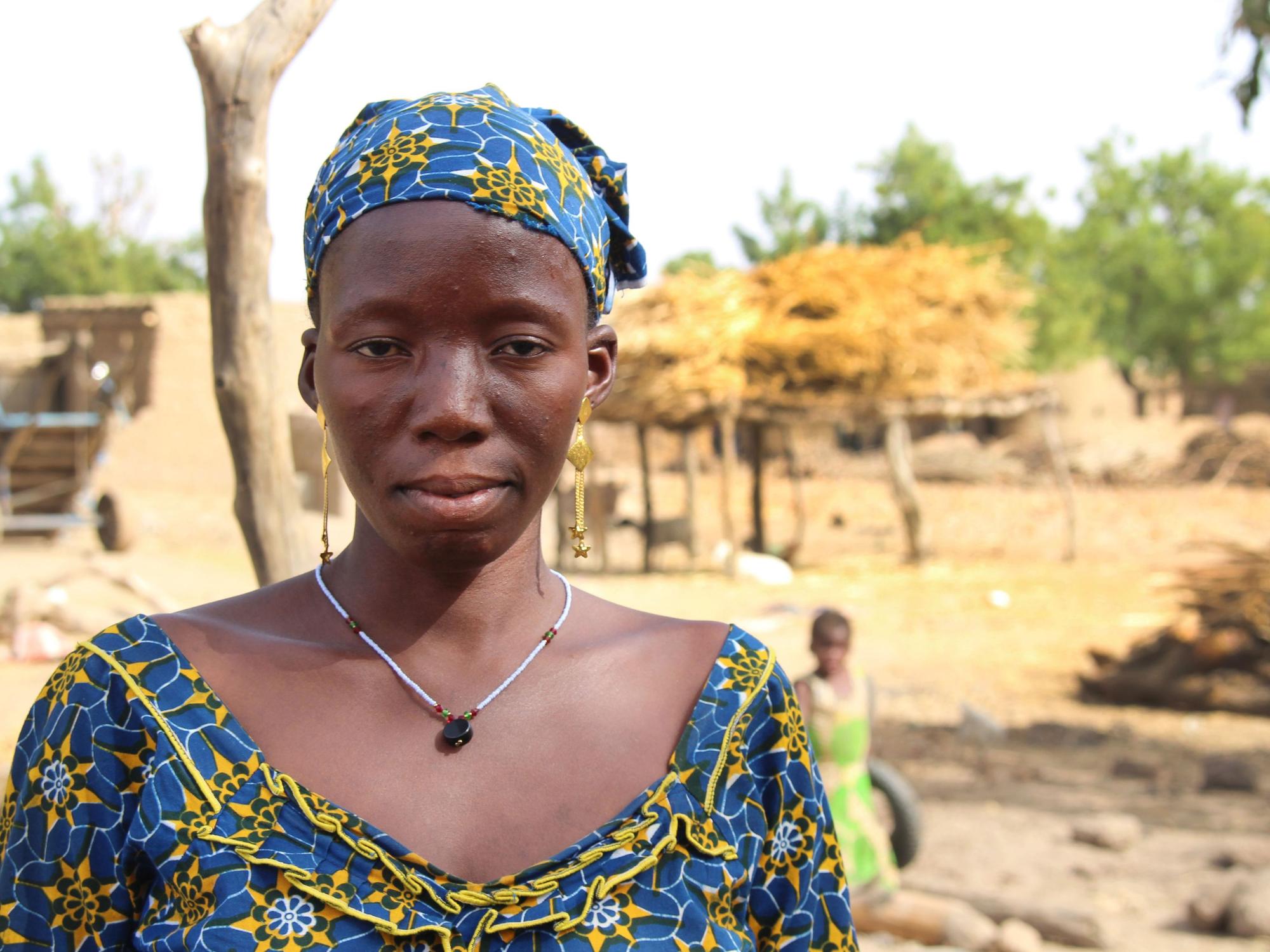 Das Bild zeigt eine junge Afrikanerin in traditioneller Kleidung vor dem Hintergrund eines Dorfes. Es handelt sich um Sanamba aus Mali, hier aufgenommen am 20. März 2018. Sie wurde schon im Alter von 14 Jahren zwangsverheiratet.