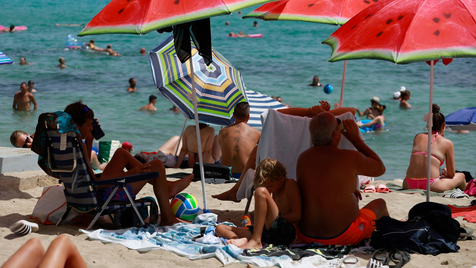 Auf dem Symbolfoto von August 2022 sieht man Strand, Meer und Menschen. Sie schwimmen, liegen in der Sonne oder unter Sonnenschirmen im Schatten.