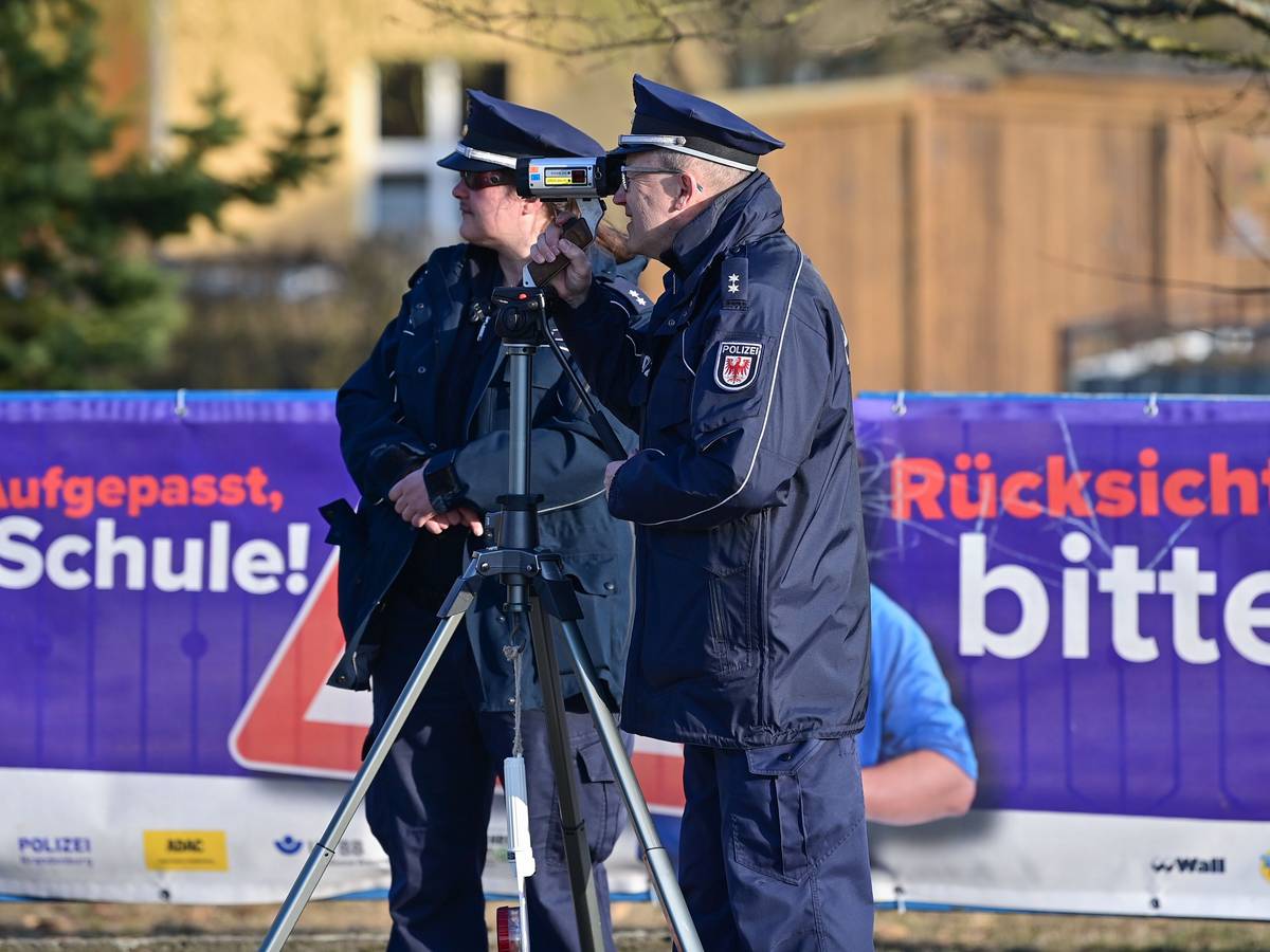 Eine Polizeioberkommissarin und ein Polizeioberkommissar stehen mit einer Laserpistole vor einer Schule und messen die Geschwindigkeit der Fahrzeuge in einer Tempo-30-Zone.