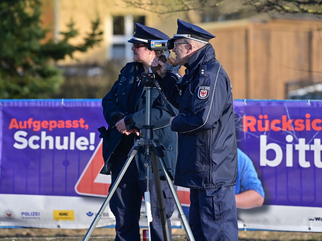 Eine Polizeioberkommissarin und ein Polizeioberkommissar stehen mit einer Laserpistole vor einer Schule und messen die Geschwindigkeit der Fahrzeuge in einer Tempo-30-Zone.