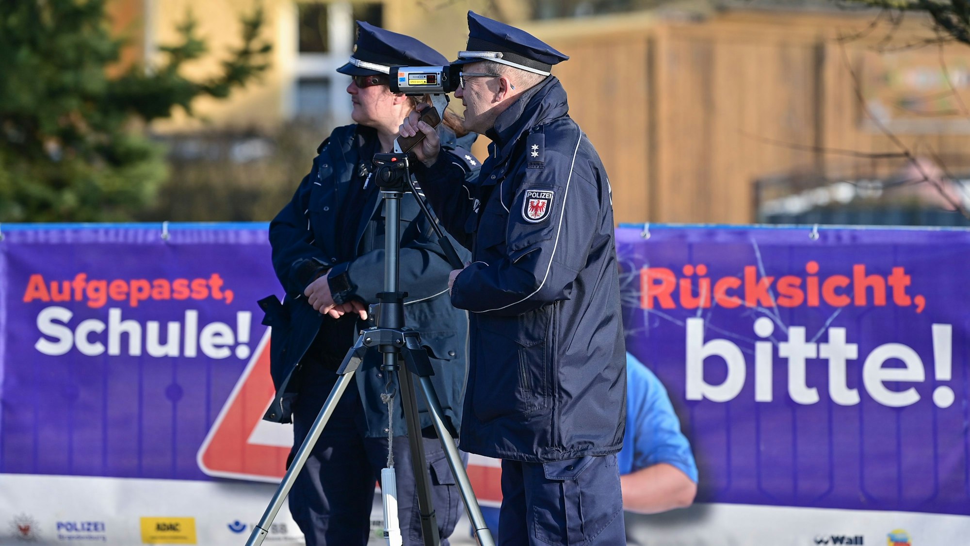 Eine Polizeioberkommissarin und ein Polizeioberkommissar stehen mit einer Laserpistole vor einer Schule und messen die Geschwindigkeit der Fahrzeuge in einer Tempo-30-Zone.